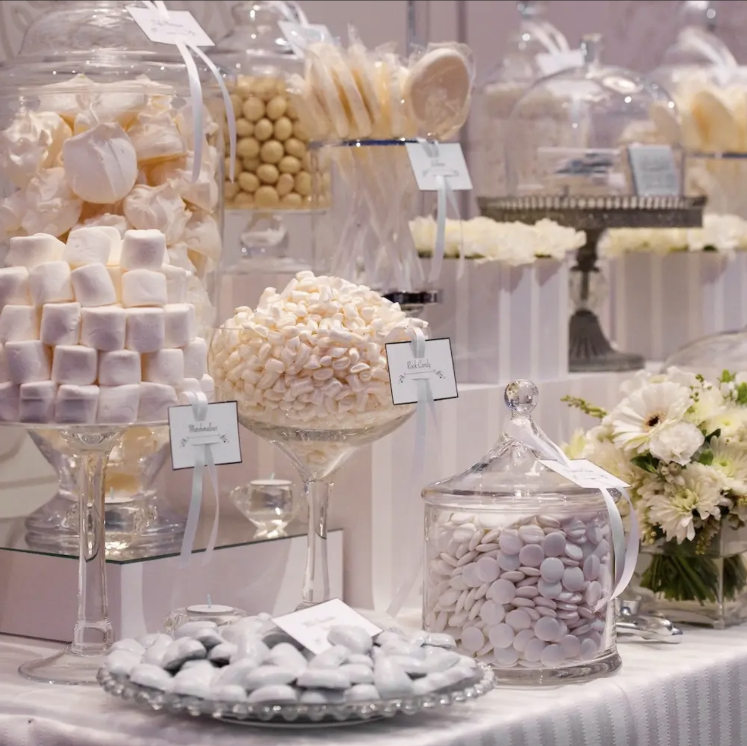 Display of glass jars and bowls filled with various white candies, marshmallows, and sweets, arranged on a table with a backdrop of flowers.