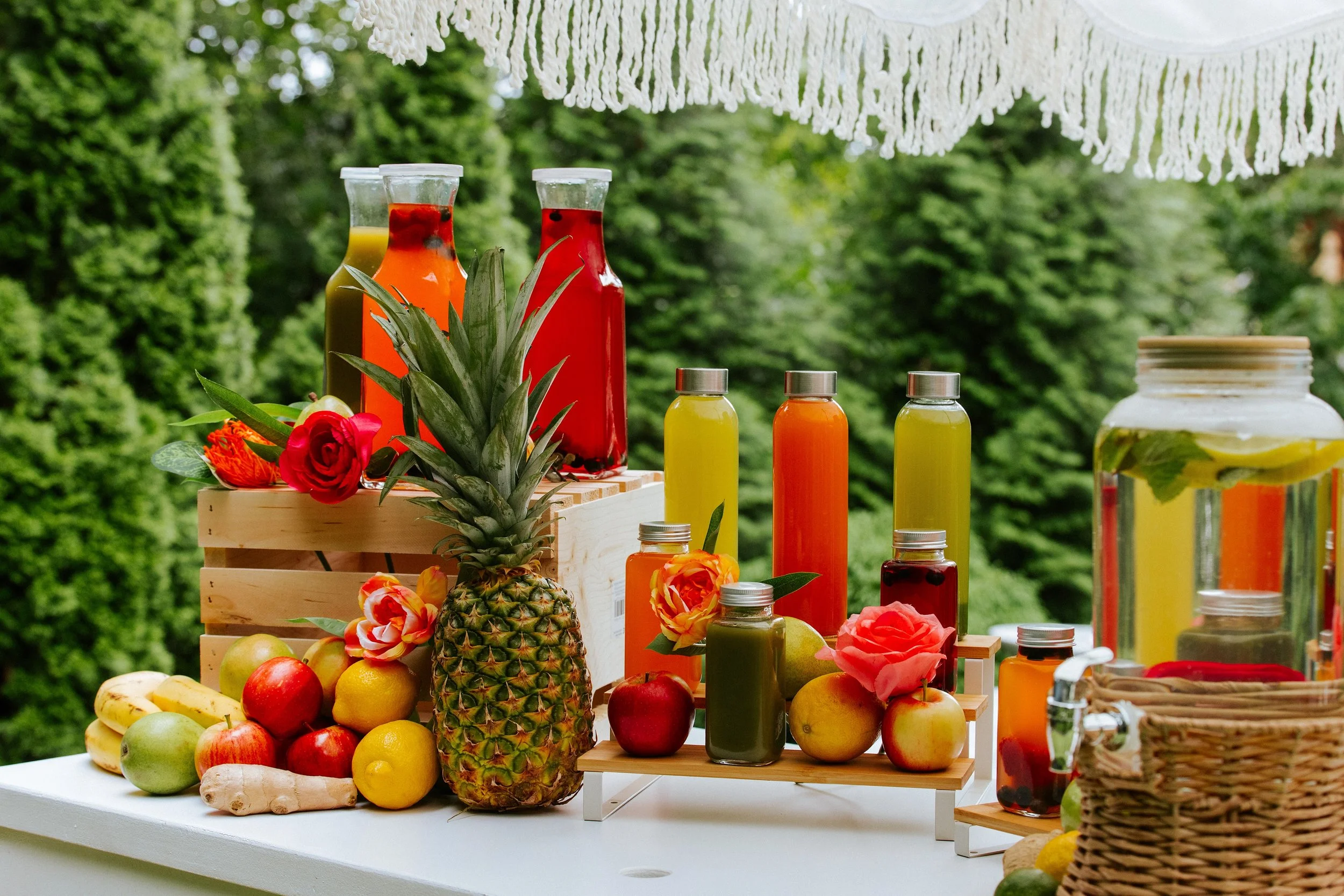 Fresh fruits like apples, bananas, oranges, pears, and pineapple with bottles of colorful juice or smoothies on a white table outdoors, decorated with a hanging white macrame.