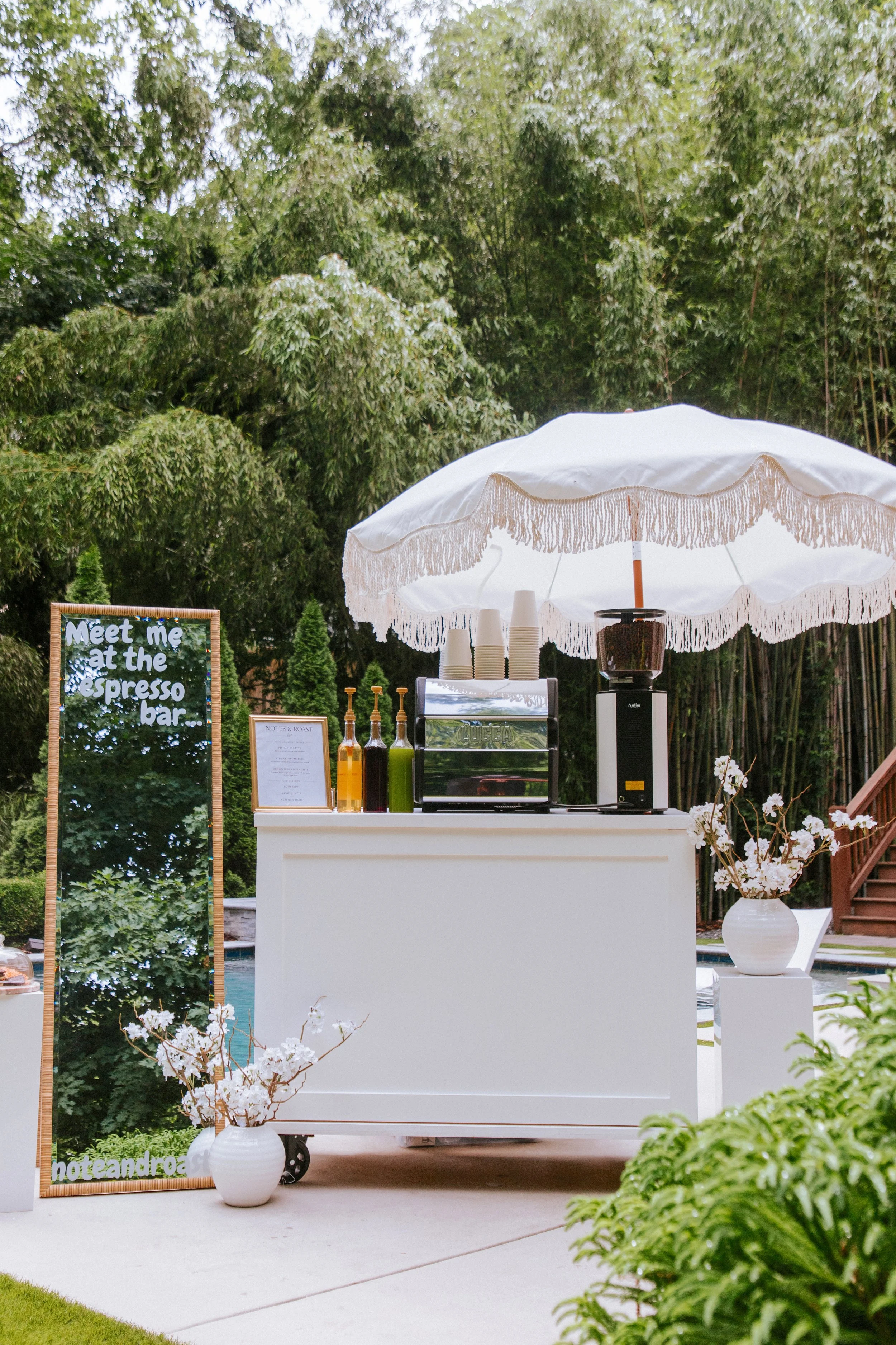 Outdoor coffee or drinks stand with a white umbrella, a white vase of flowers, and a sign that says 'Meet me at the espresso bar.' There are bottles, cups, and a coffee machine on the stand, surrounded by trees and greenery.