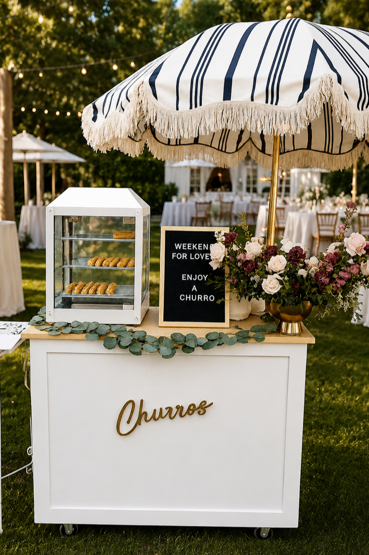 A churro stand at an outdoor event decorated with a striped umbrella, a vase of pink and white flowers, a sign that says "Weekend for Love, Enjoy a Churro," and a glass display case with churros inside. In the background, there are tables with white tablecloths and string lights.