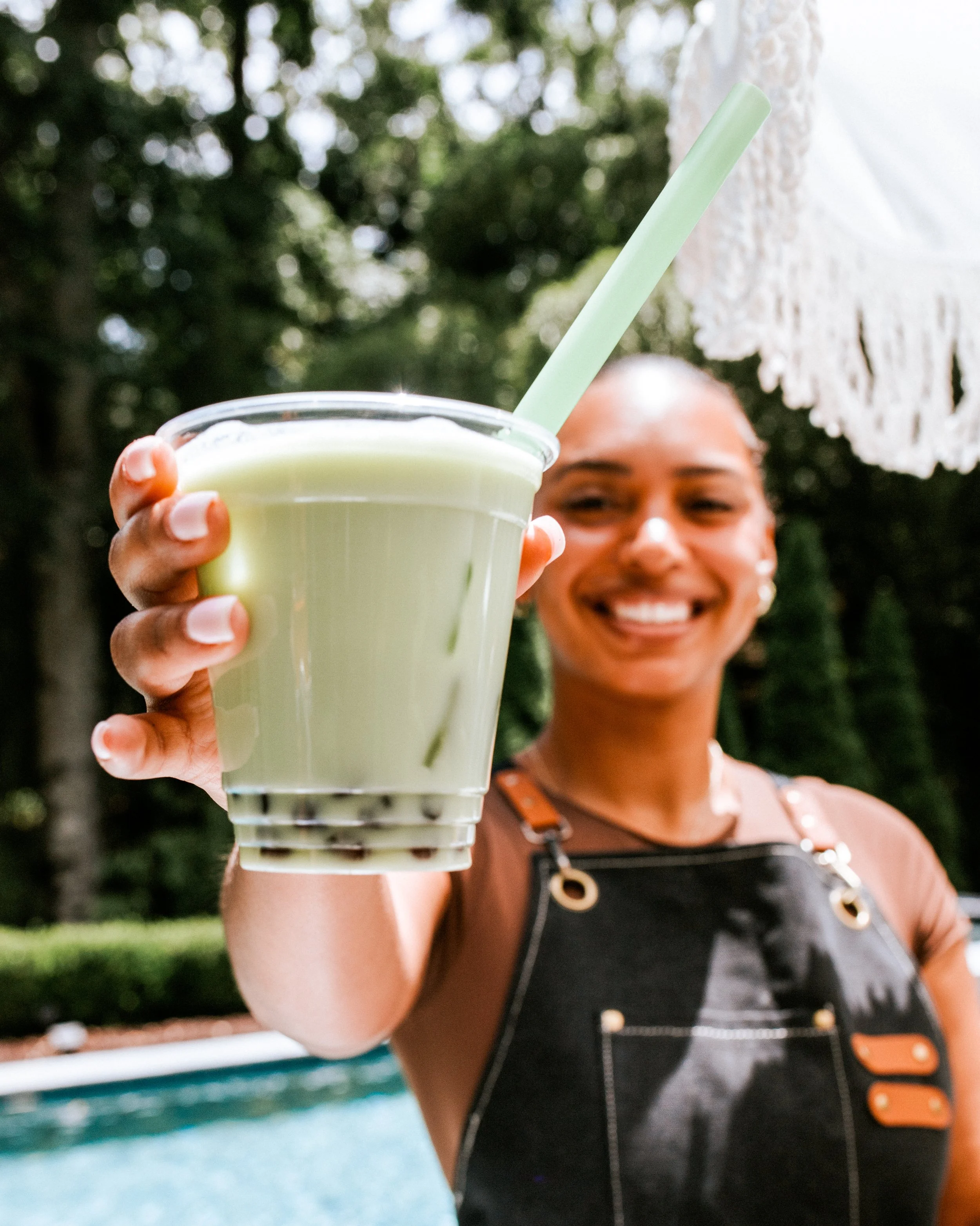 A woman smiling and holding a glass of pale green beverage with a straw outdoors, with trees and a white decorative hanging in the background.
