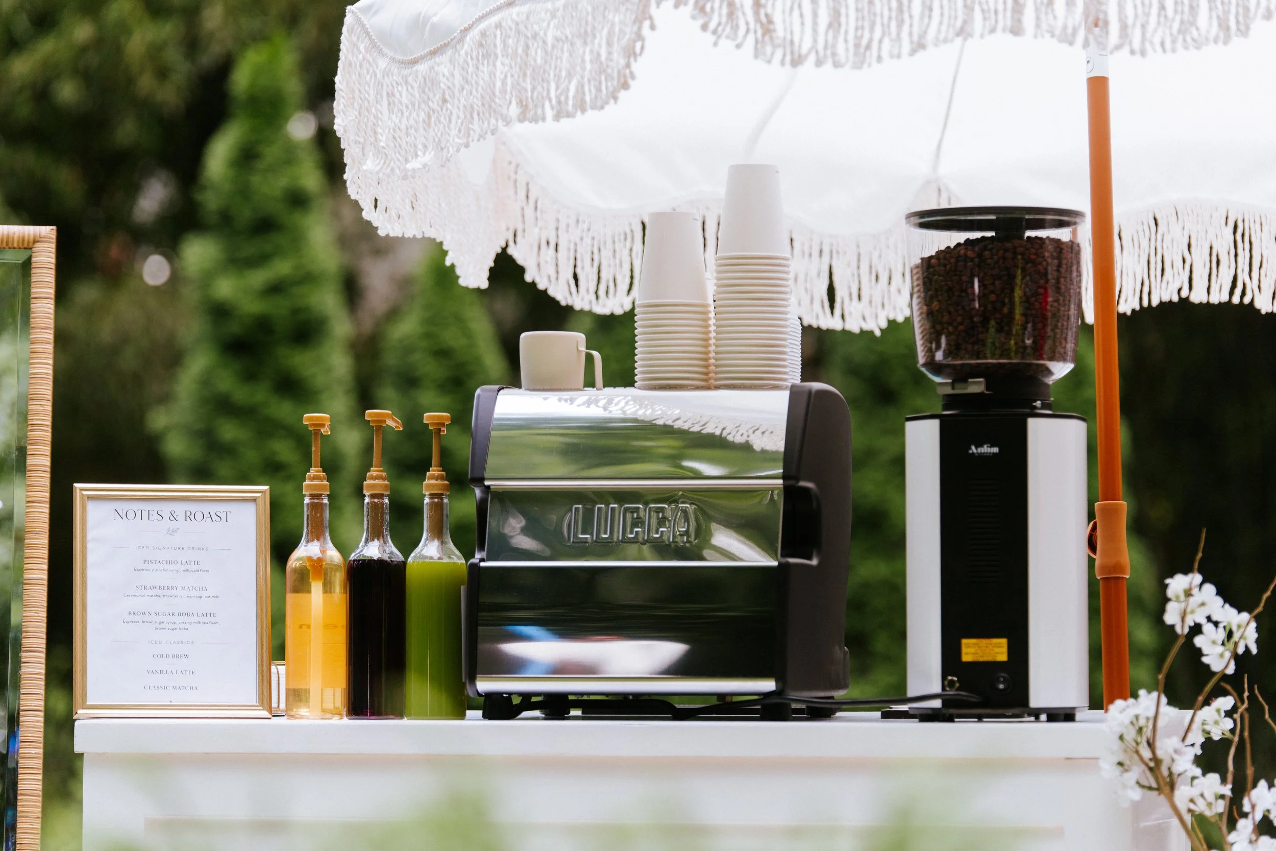 A coffee stand with a white lace canopy, plastic cups, syrup bottles, a coffee machine, and a menu sign, outdoors in a park or garden setting.