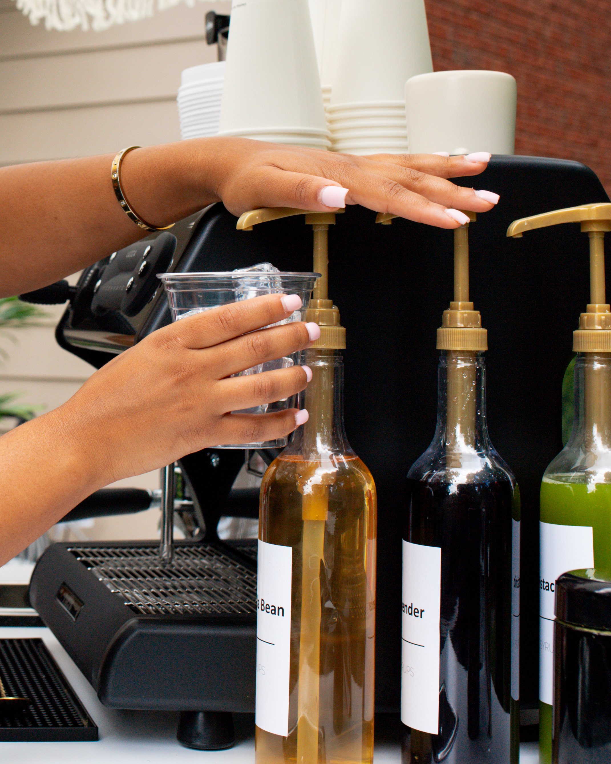A hand with white painted nails and a gold bracelet is pressing a pump on a clear bottle of liquid, with other bottles with labels and a black device visible in the background.