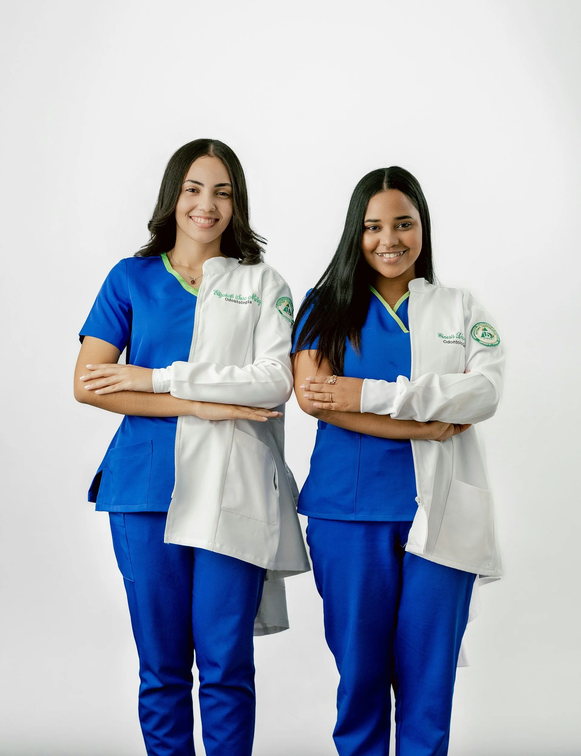 Two women in dental uniforms, standing against a white background, smiling with arms crossed.