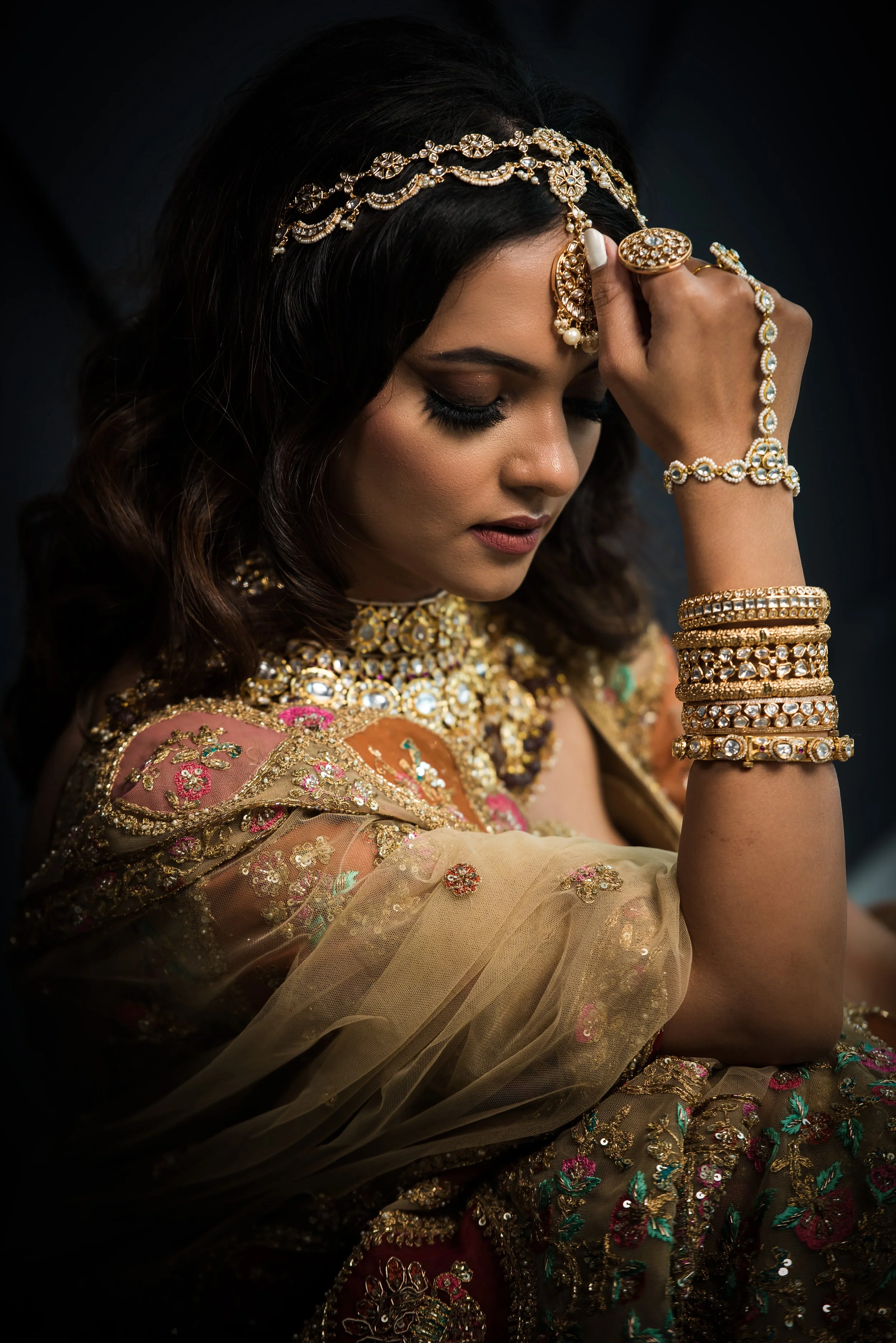 Indian bride in an intimate portrait wearing kundan and gold bridal jewelry with layered bangles and headpiece.