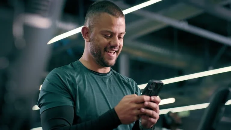 A man smiling while looking at his smartphone in a gym.