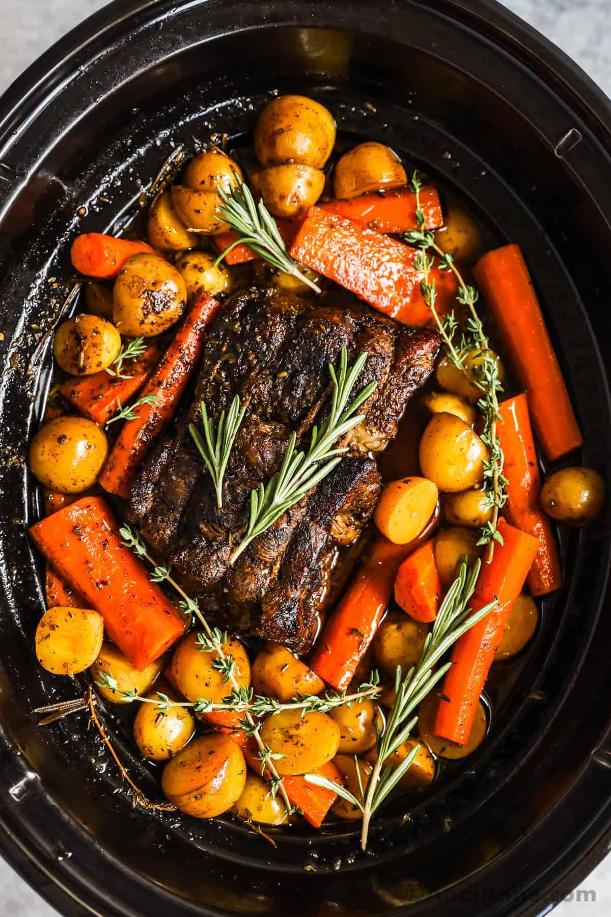 Cooked beef roast with baby potatoes, carrots, and sprigs of fresh rosemary and thyme in a slow cooker.