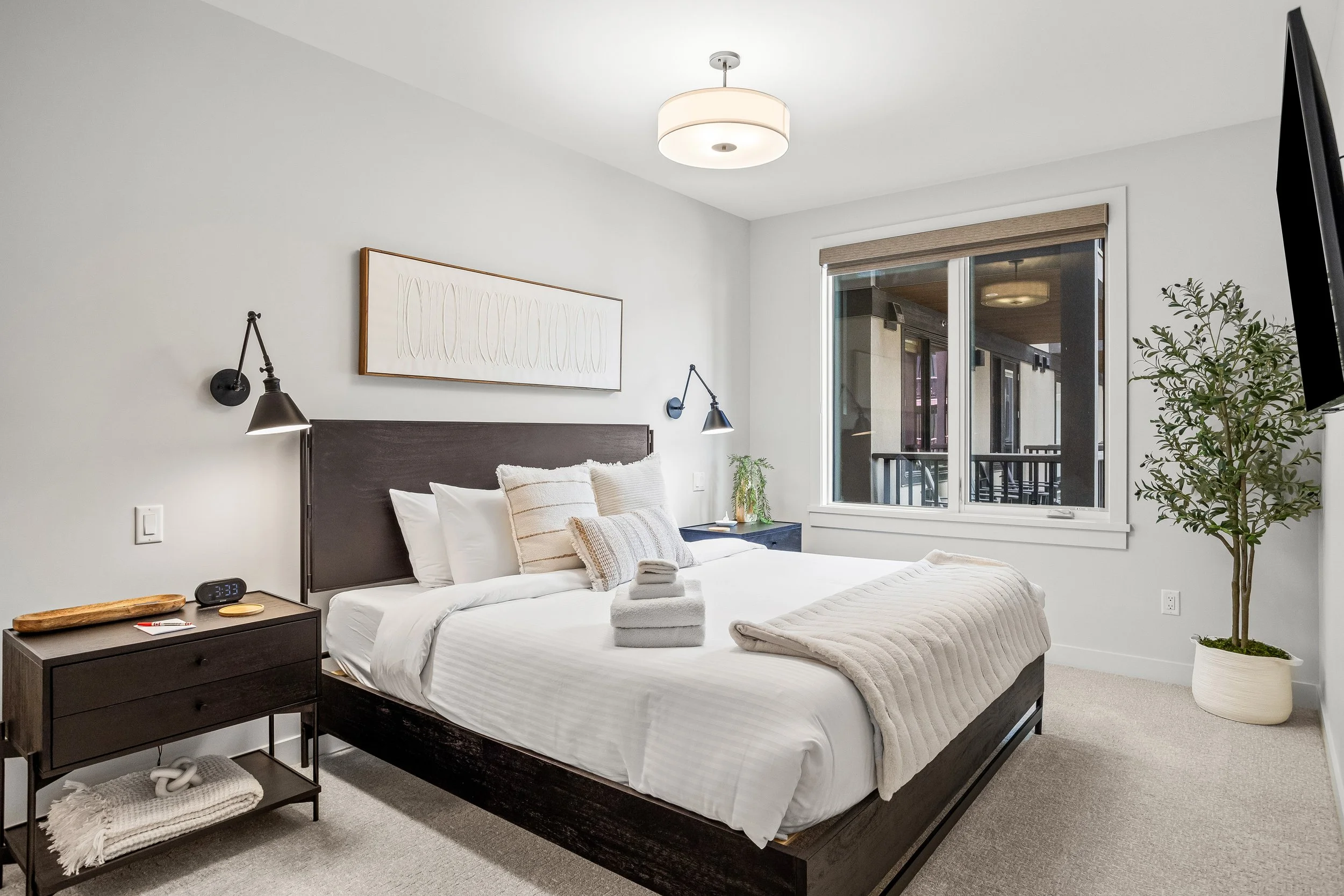 A modern bedroom with a dark wooden bed frame, white bedding, and decorative pillows. Nightstands on either side with black wall-mounted lamps, a window with a view of a balcony, and a potted plant near the corner.