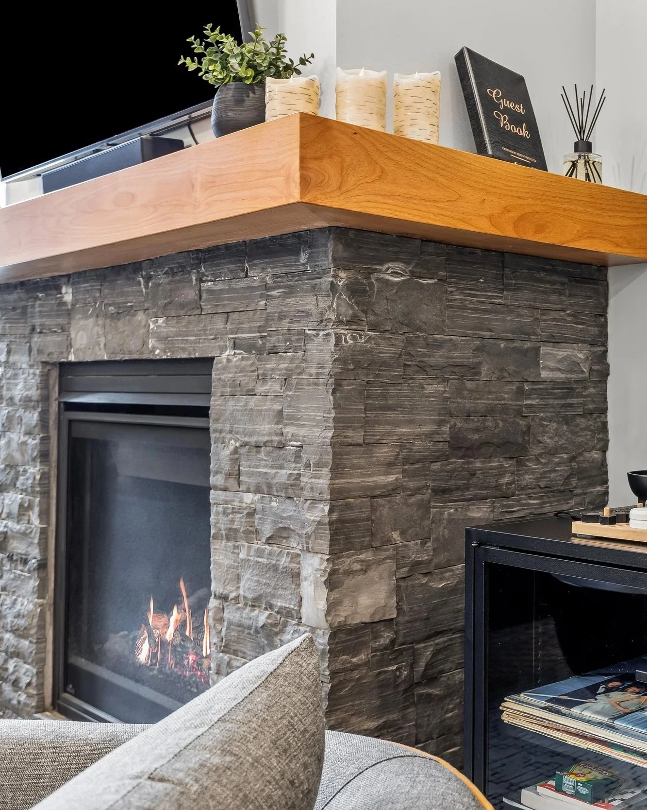 Close-up of a modern living room corner featuring a stone fireplace with a wooden mantel, decorated with a plant, a guest book, and a diffuser. Part of a gray sofa with a cushion and a black cabinet with magazines are also visible.