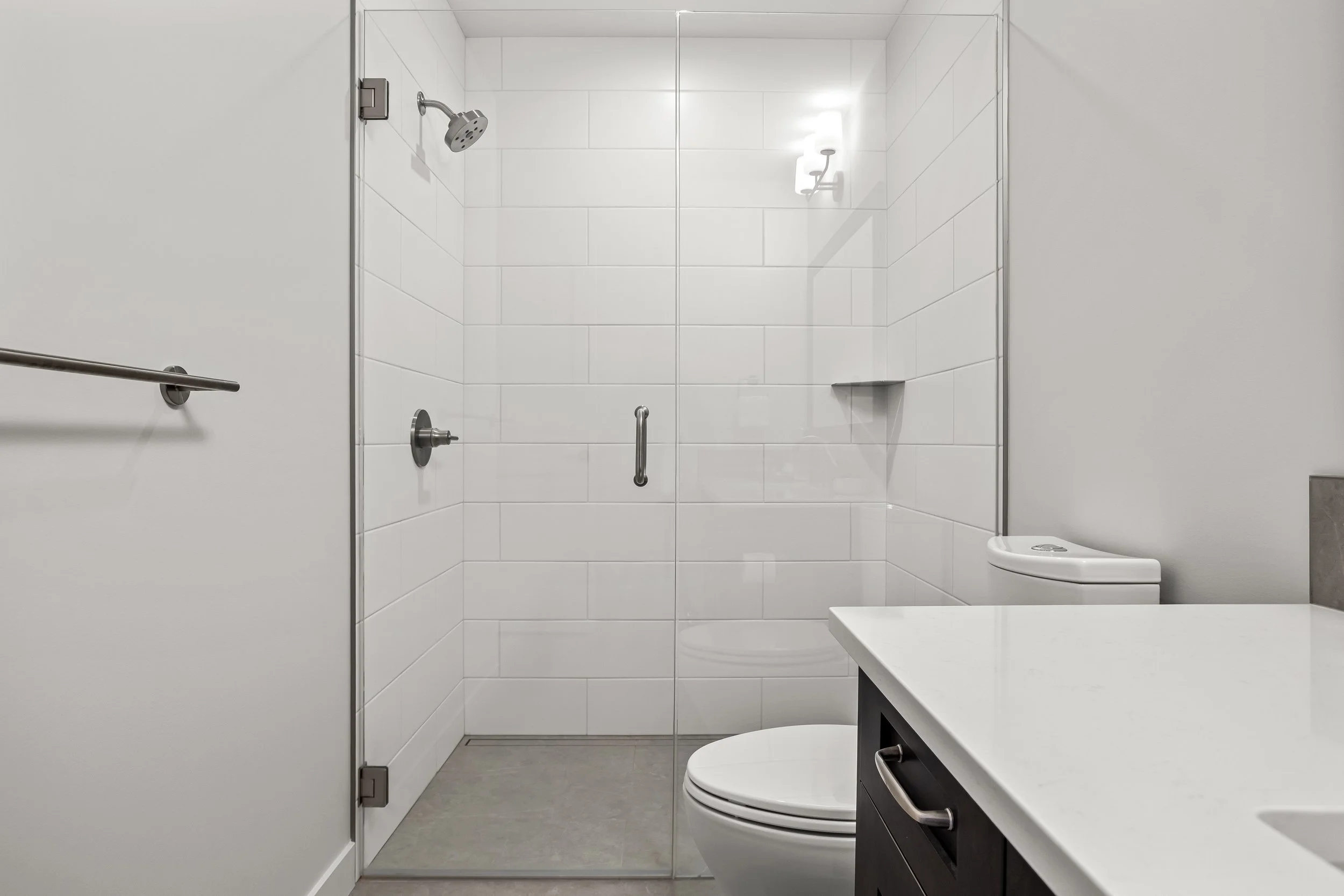 Modern bathroom with a glass-enclosed shower, white tiled walls, a white toilet, and a black vanity with a white countertop.