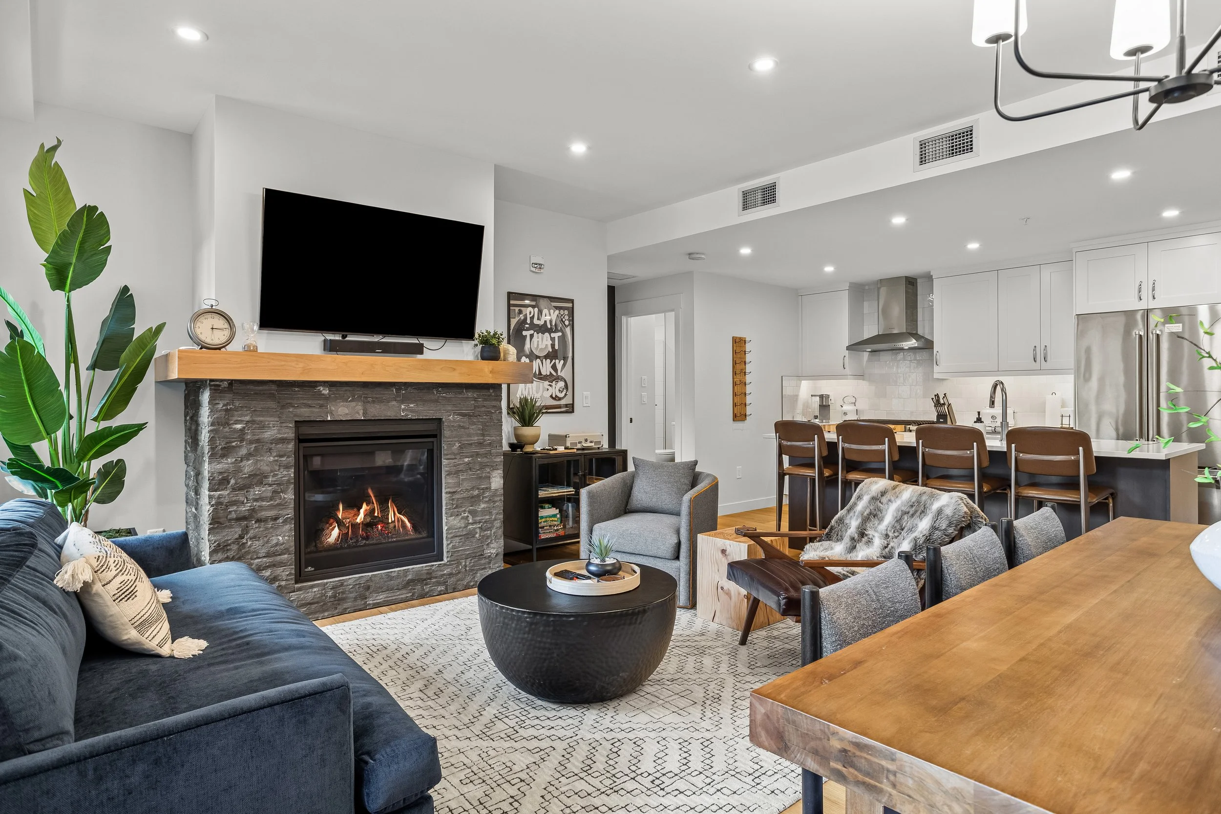 Modern, open-concept living room with a stone fireplace, mounted flat-screen TV, gray sofas, a black round coffee table, potted plants, and a view into the Kitchen with a bar counter and stools.