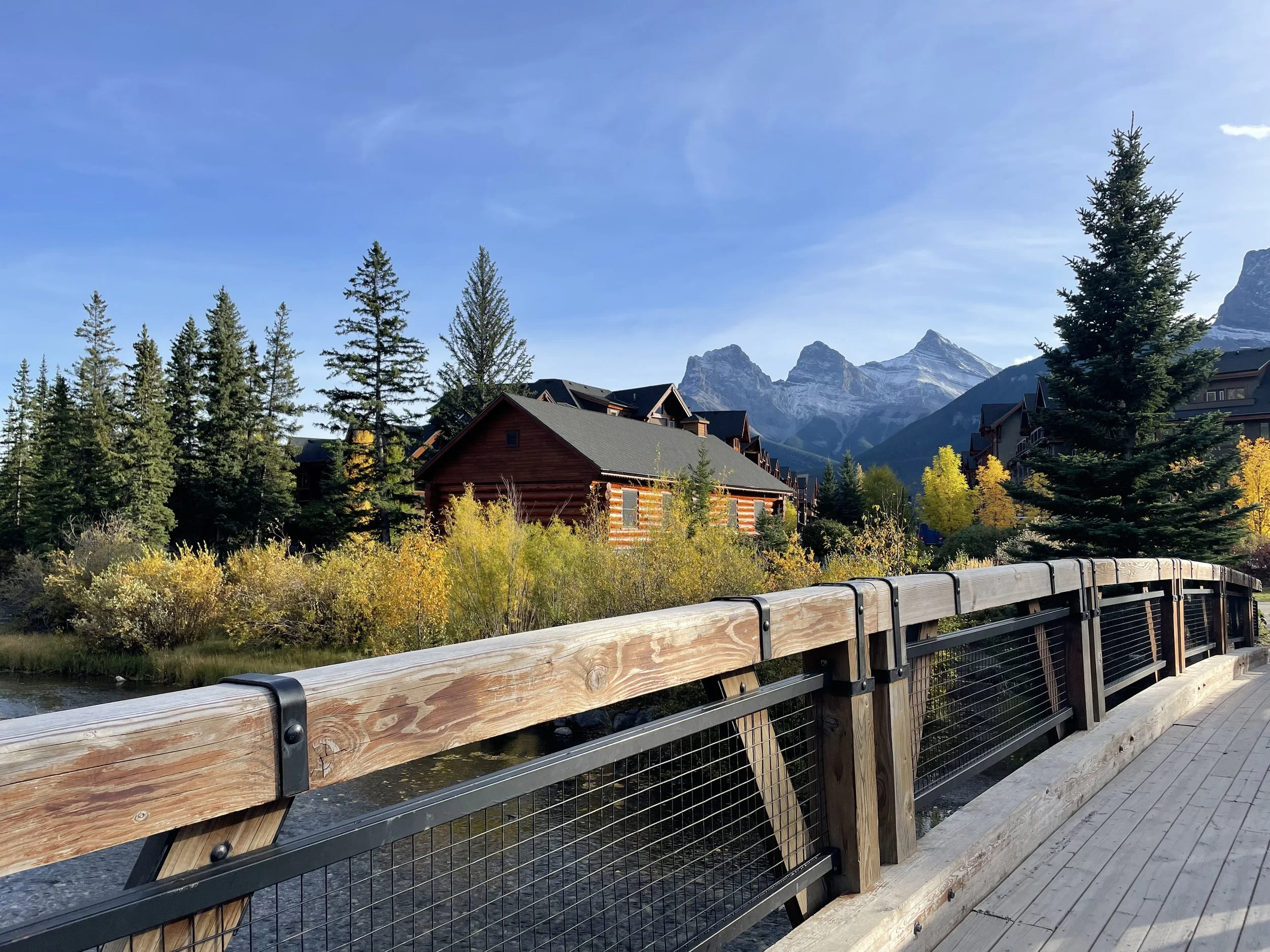Scene of a wooden bridge in Canmore, Alberta, overlooking a river with forested trees, a red cabin, and mountains in the background under a clear blue sky.