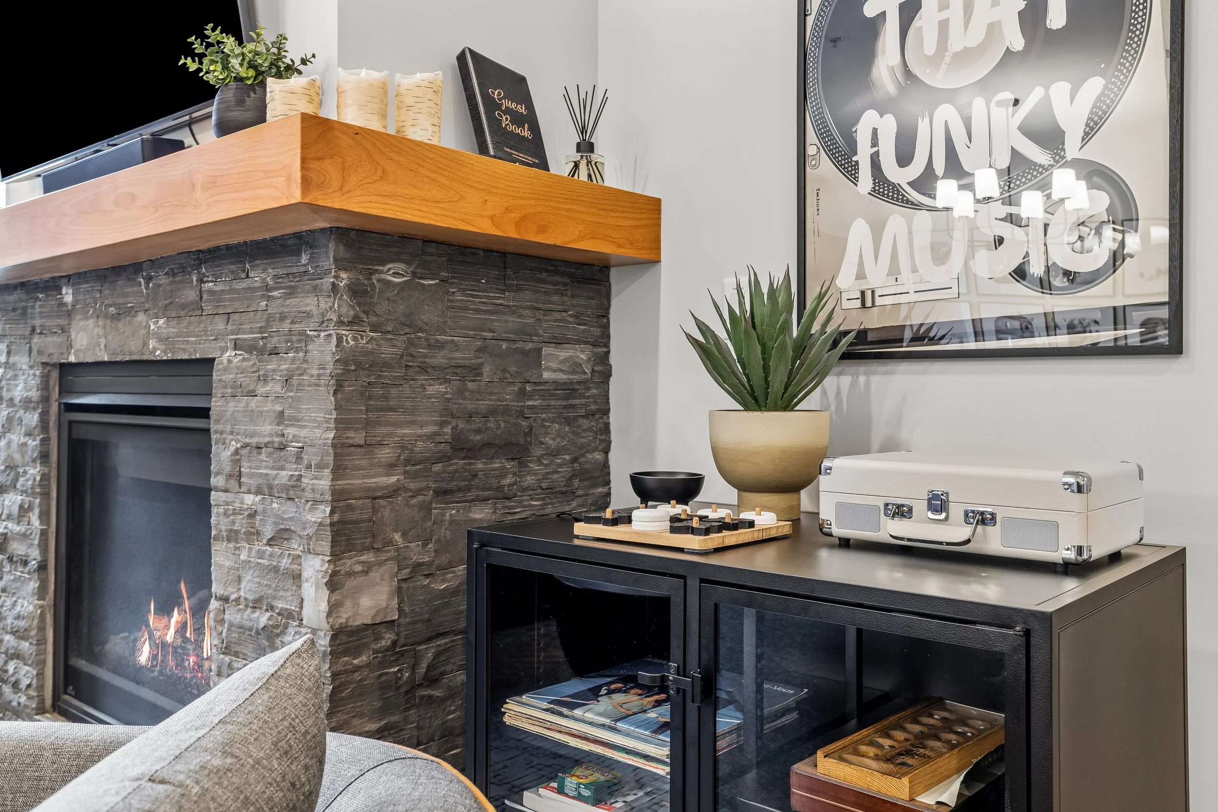 Interior of a living room with a stone fireplace, a black cabinet topped with a potted plant, vinyl records, and a vintage box, a large framed art print on the wall reading 'That Funky Music,' and decorative objects on the mantle including a guest bo