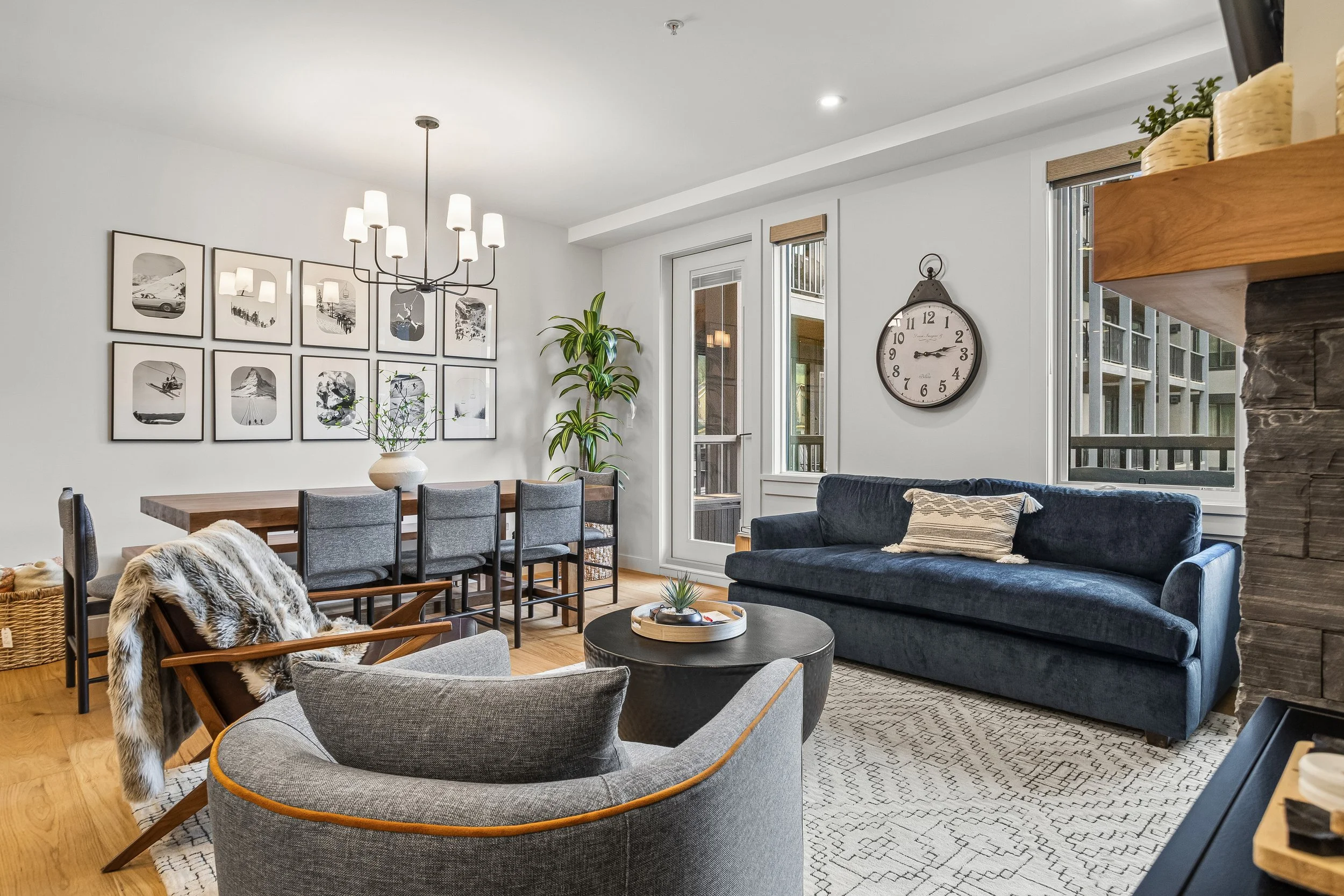 A modern living room with a dining area, gray and navy furniture, a wall clock, and large windows letting in natural light.