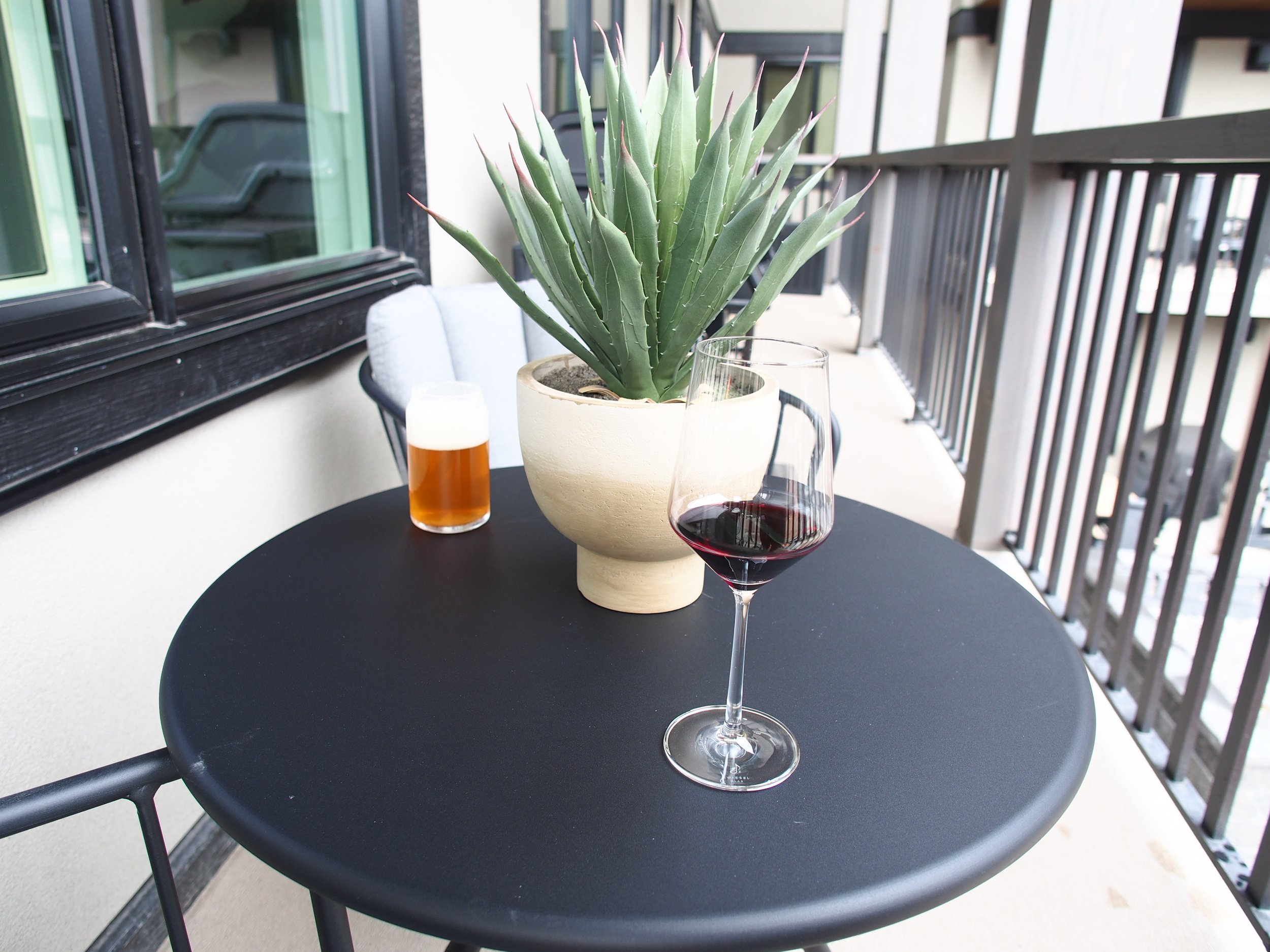 A black round table on a balcony with a large potted succulent, a glass of red wine, and a glass of beer.