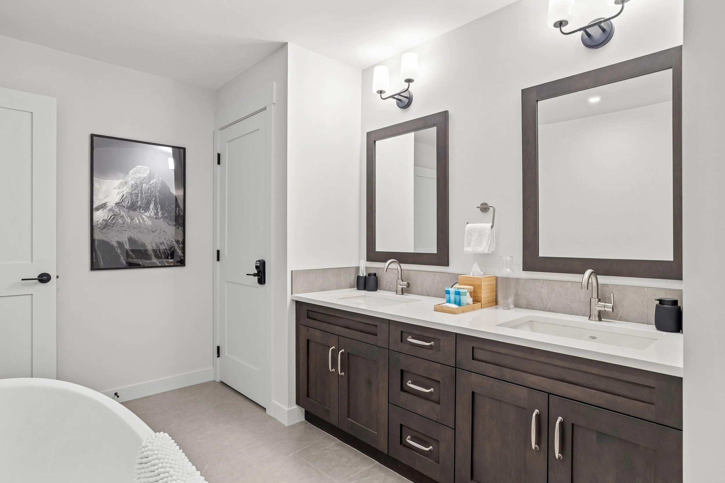 Modern bathroom with double vanity, two mirrors, wall-mounted light fixtures, and a closed white door. Wall art depicts a mountain scene.