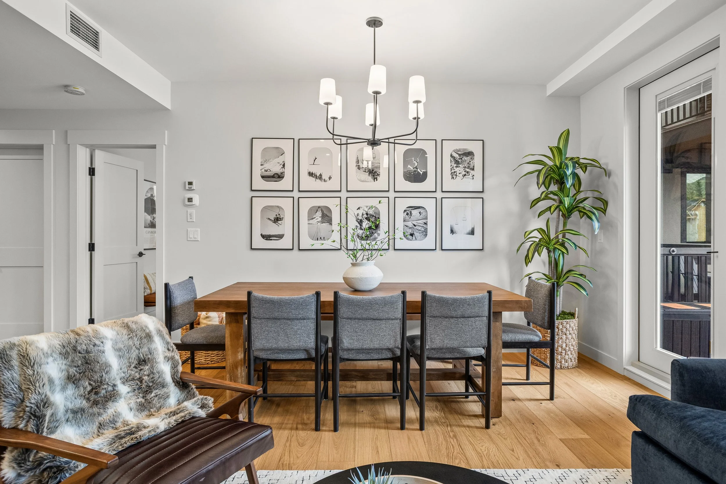 Modern dining room with a wooden dining table surrounded by six gray chairs. A black and white photo art gallery hangs on the white wall. A large potted plant is near a glass door leading outside. A chandelier with white shades hangs above the table.