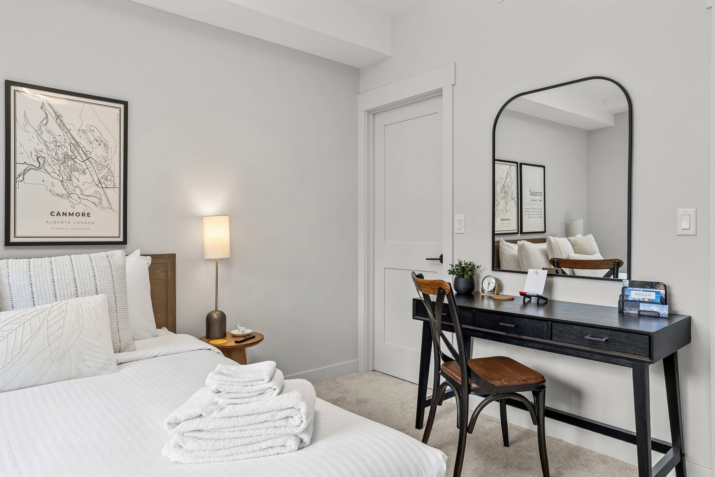 Canmore condo bedroom with white bed, bedside table, lamp, wall art, black desk with mirror, chairs, and decor.