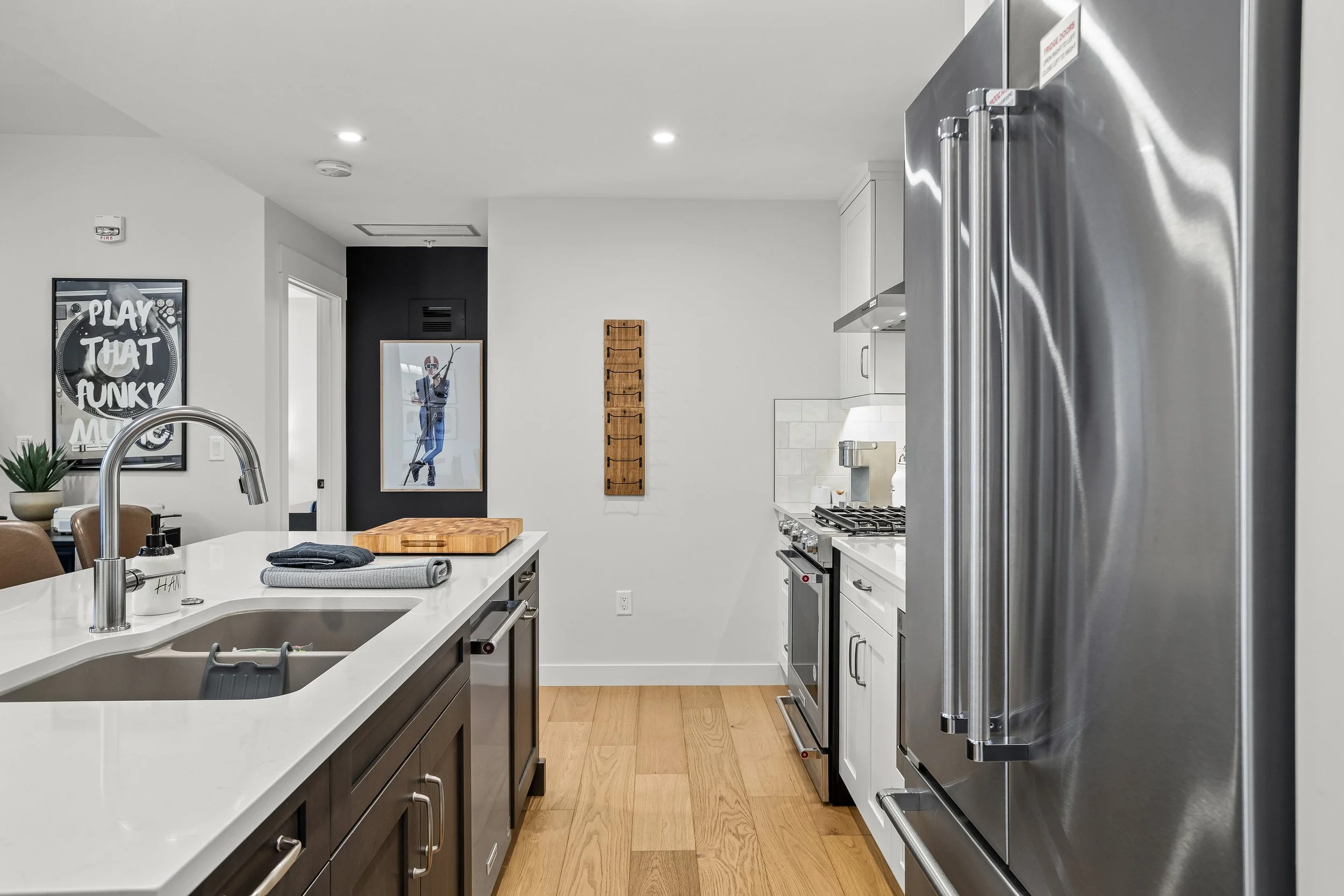 Modern kitchen with white counters, stainless steel appliances, and wood flooring, decorated with wall art and a small wooden rack on the wall.