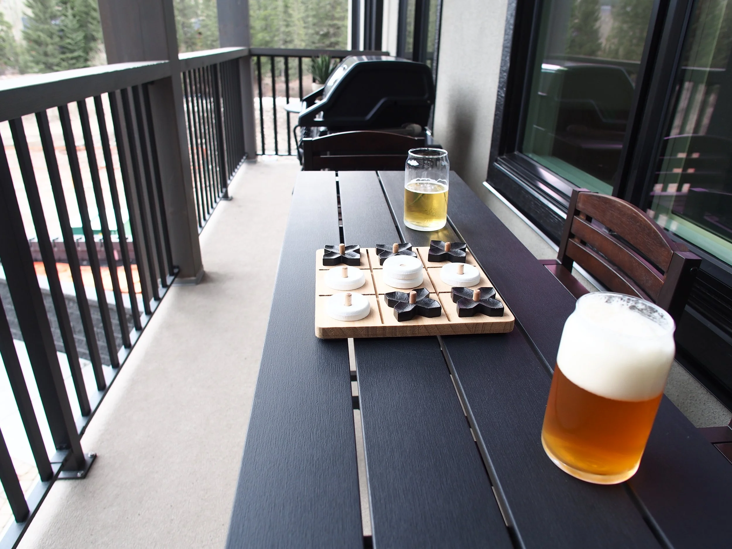 On a black outdoor table, there are two glasses of beer, a wooden tic-tac-toe game with black and white pieces, and a tray with some playing pieces, on a balcony with a railing and trees in the background.
