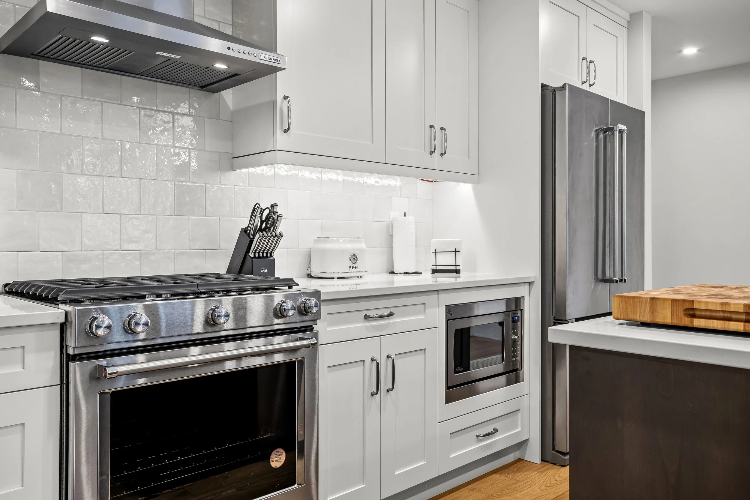 Modern kitchen with white cabinets, stainless steel appliances including a stove, microwave, and refrigerator, and a wooden cutting board on a kitchen island.