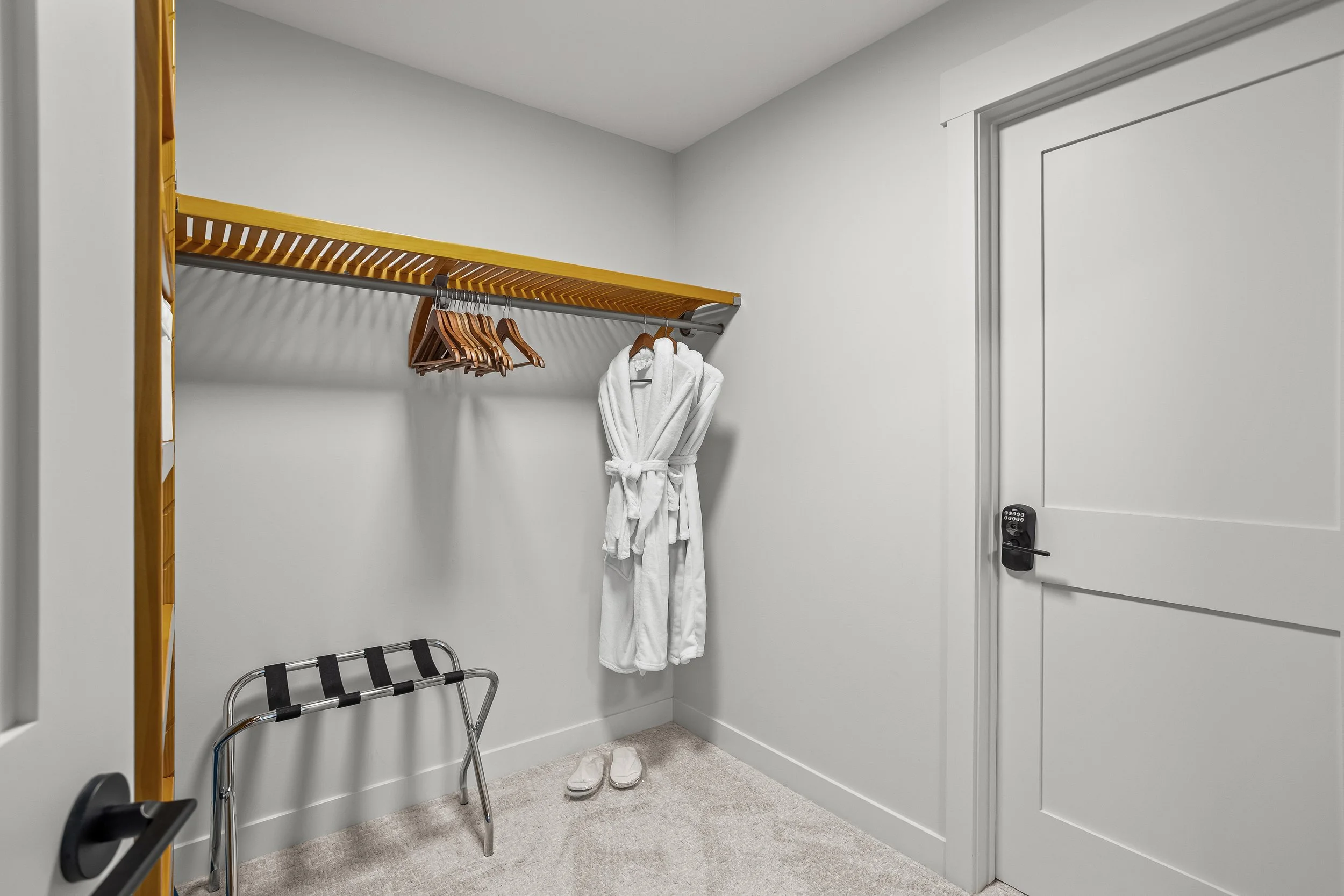 Empty walk-in closet with a hanging rod, a yellow shelf, a white bathrobe hanging, a pair of slippers on the beige carpet, and a small black luggage rack.