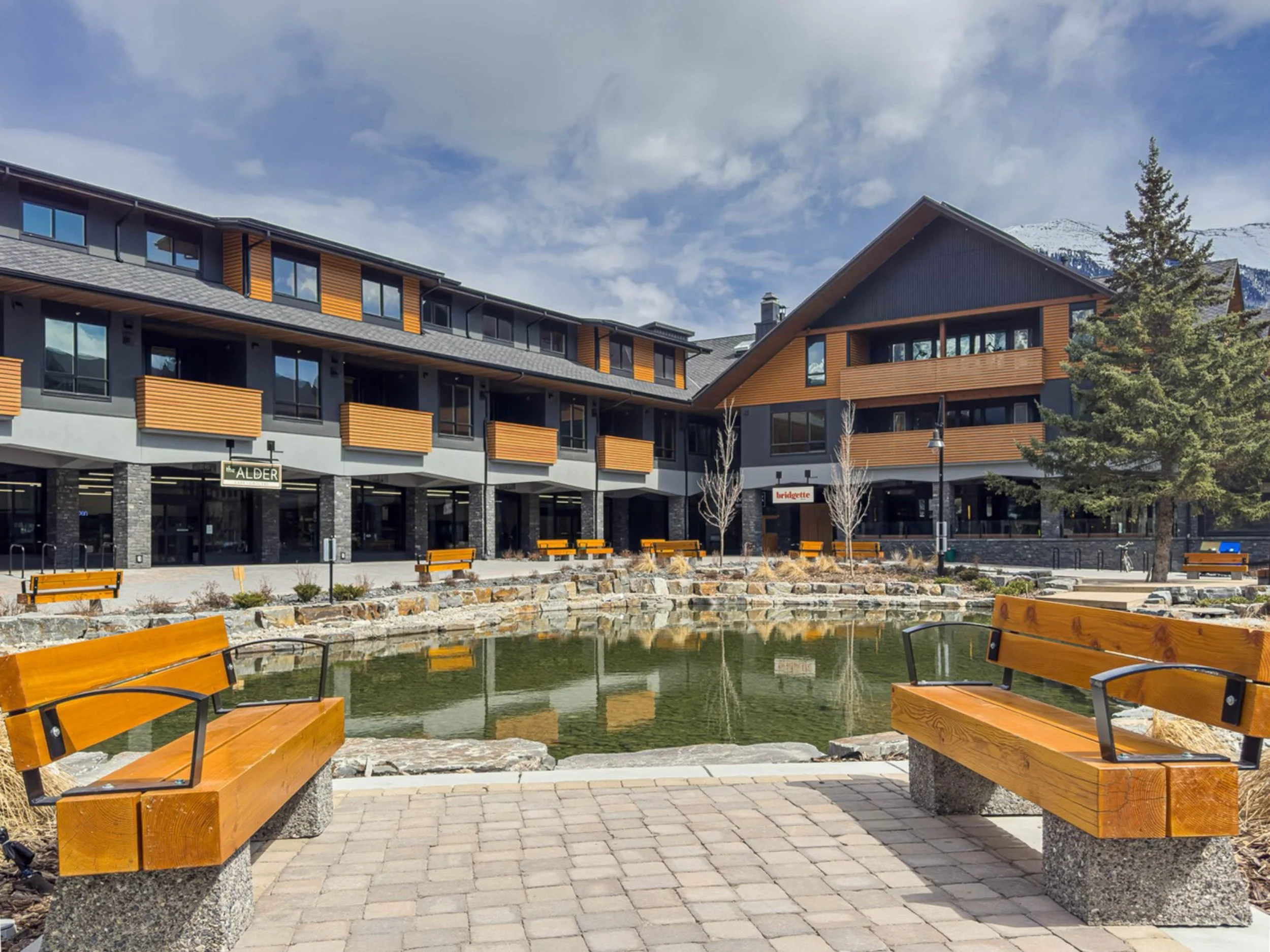 Modern apartment complex with a pond and benches in the foreground, trees, and mountains in the background.