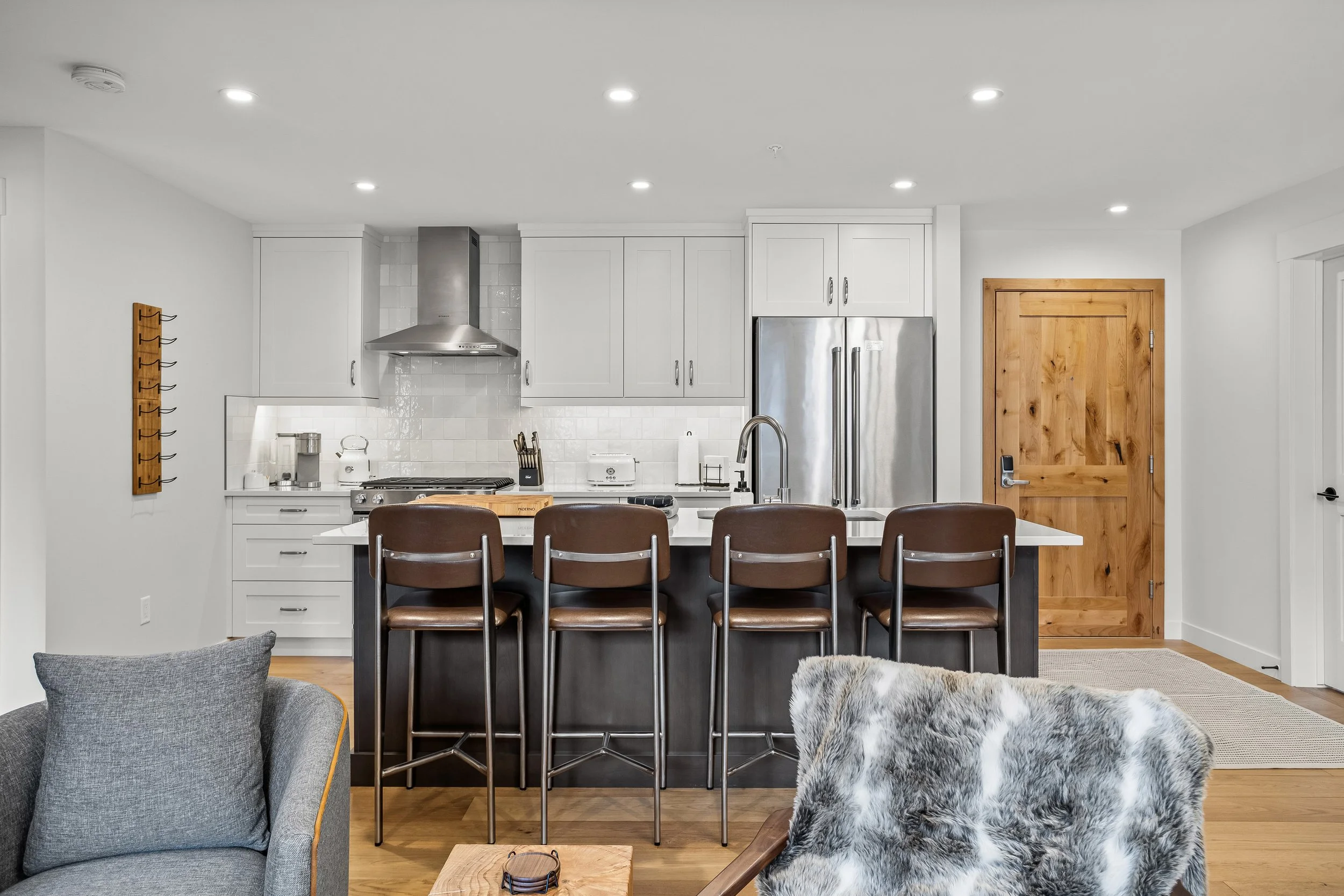 Modern kitchen with white cabinets, stainless steel refrigerator, wooden doorway, bar stools, and a living area with a gray armchair and fur-covered chair.