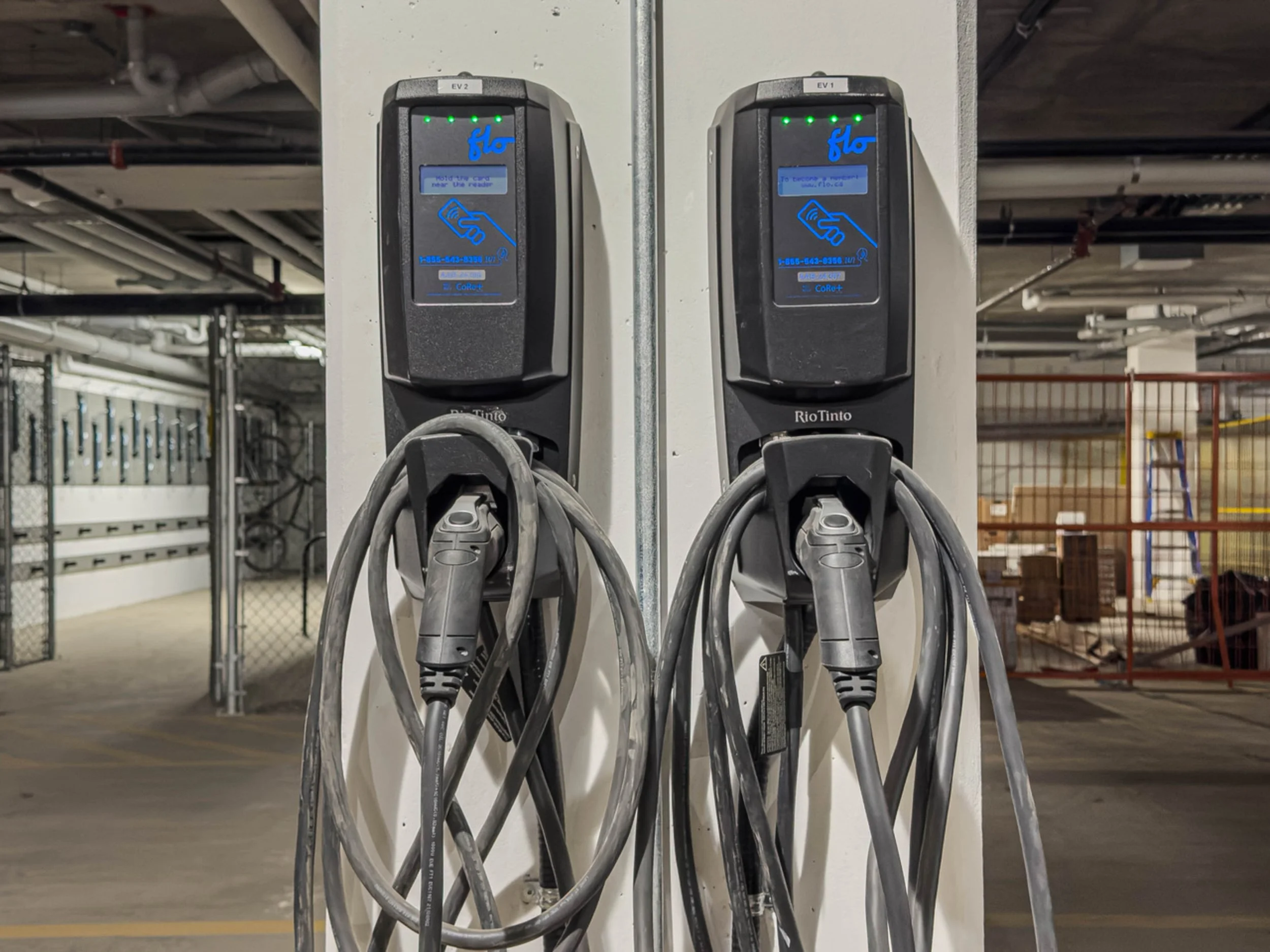 Two electric vehicle charging stations with cables hanging, in an indoor parking garage with parked bikes and construction area in the background.