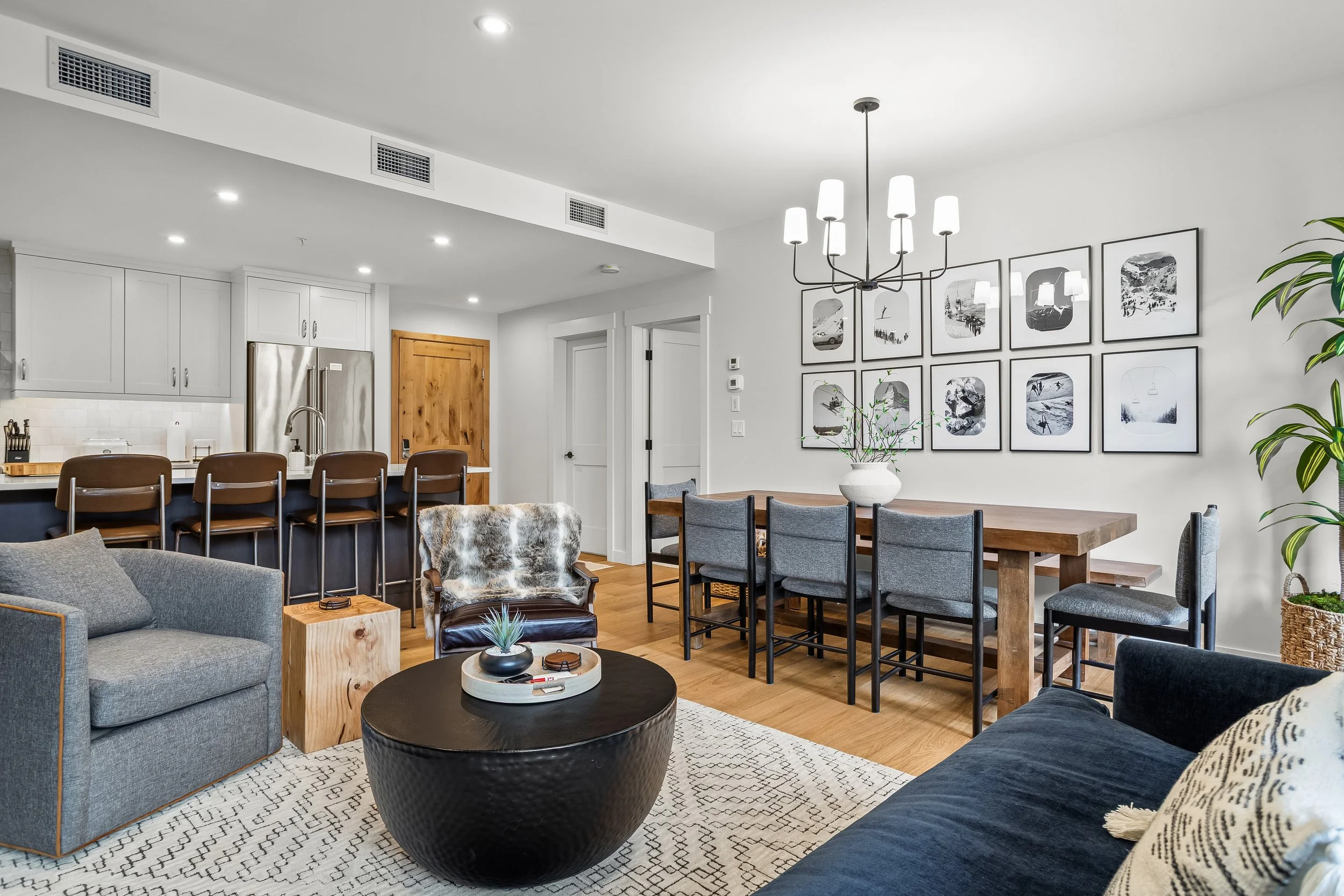 Modern open-concept living room and kitchen with gray sofa, armchair, black round coffee table, wooden dining table with six gray chairs, white walls, framed black-and-white photographs, and a white vase with green plants