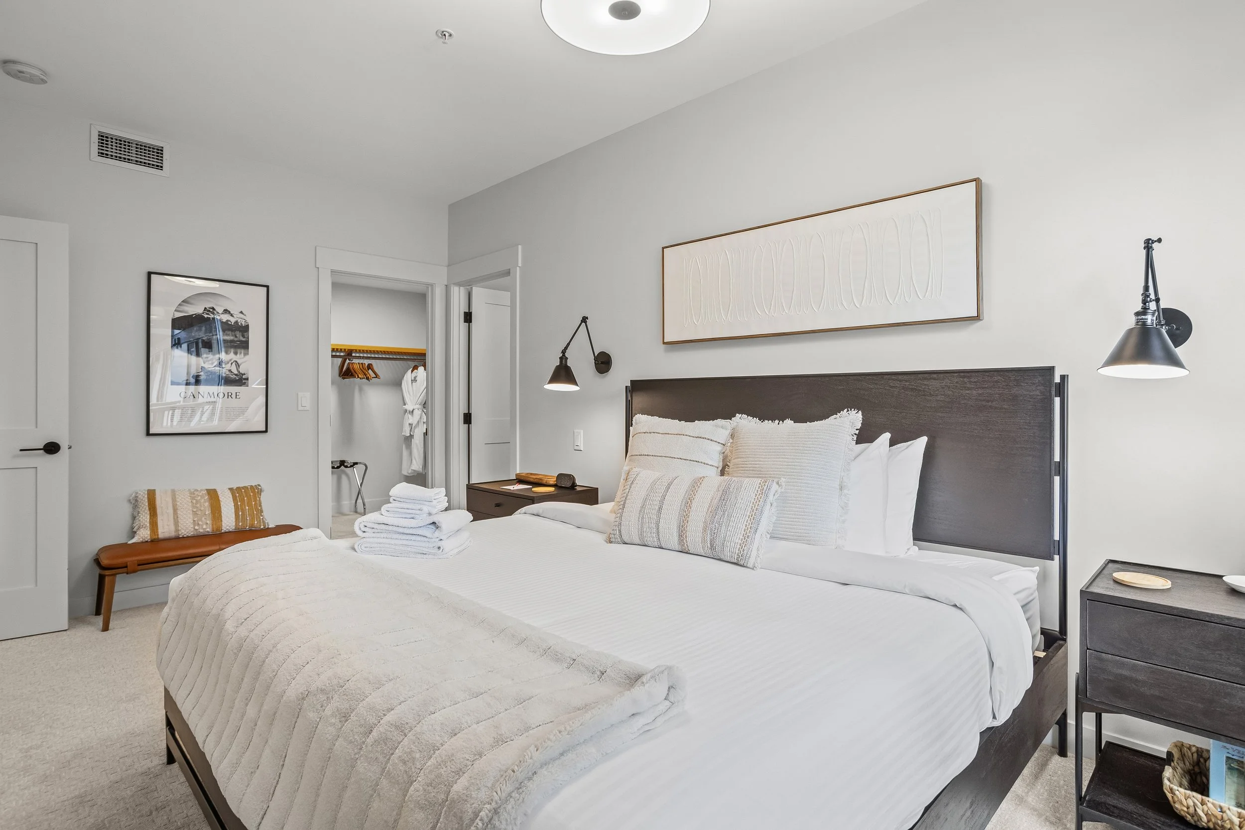 Modern bedroom with a large bed featuring white linens and a beige throw, dark wood headboard, and matching nightstand. There are several decorative pillows on the bed, and picture frames on the walls. An open closet with robes and hangers is visible