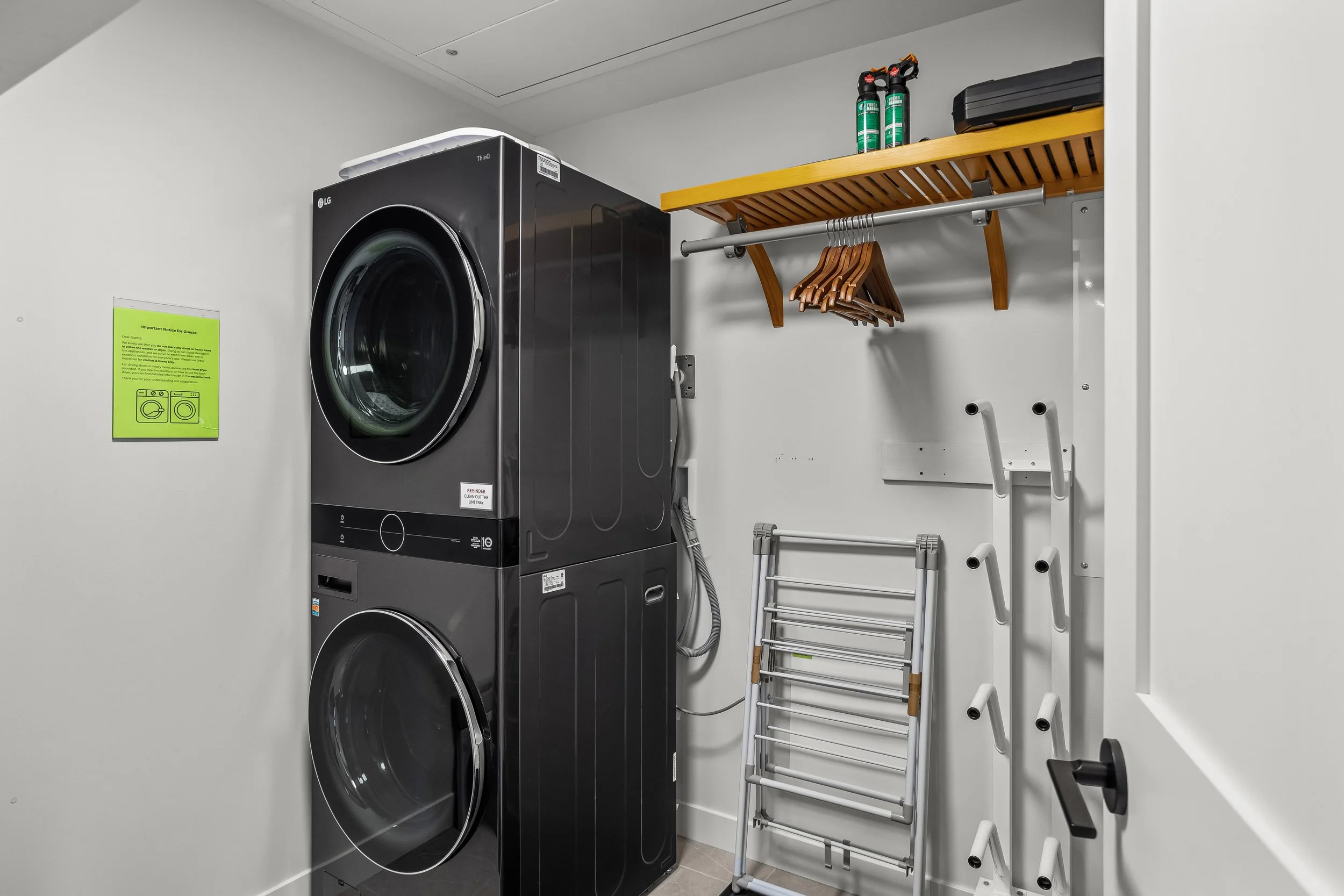 A laundry room features a black LG front-loading washing machine and dryer stacked vertically, with white walls. To the right, a wooden shelf holds two bottles of cleaning spray, and below the shelf, a metal rod with hangers. A foldable drying rack l
