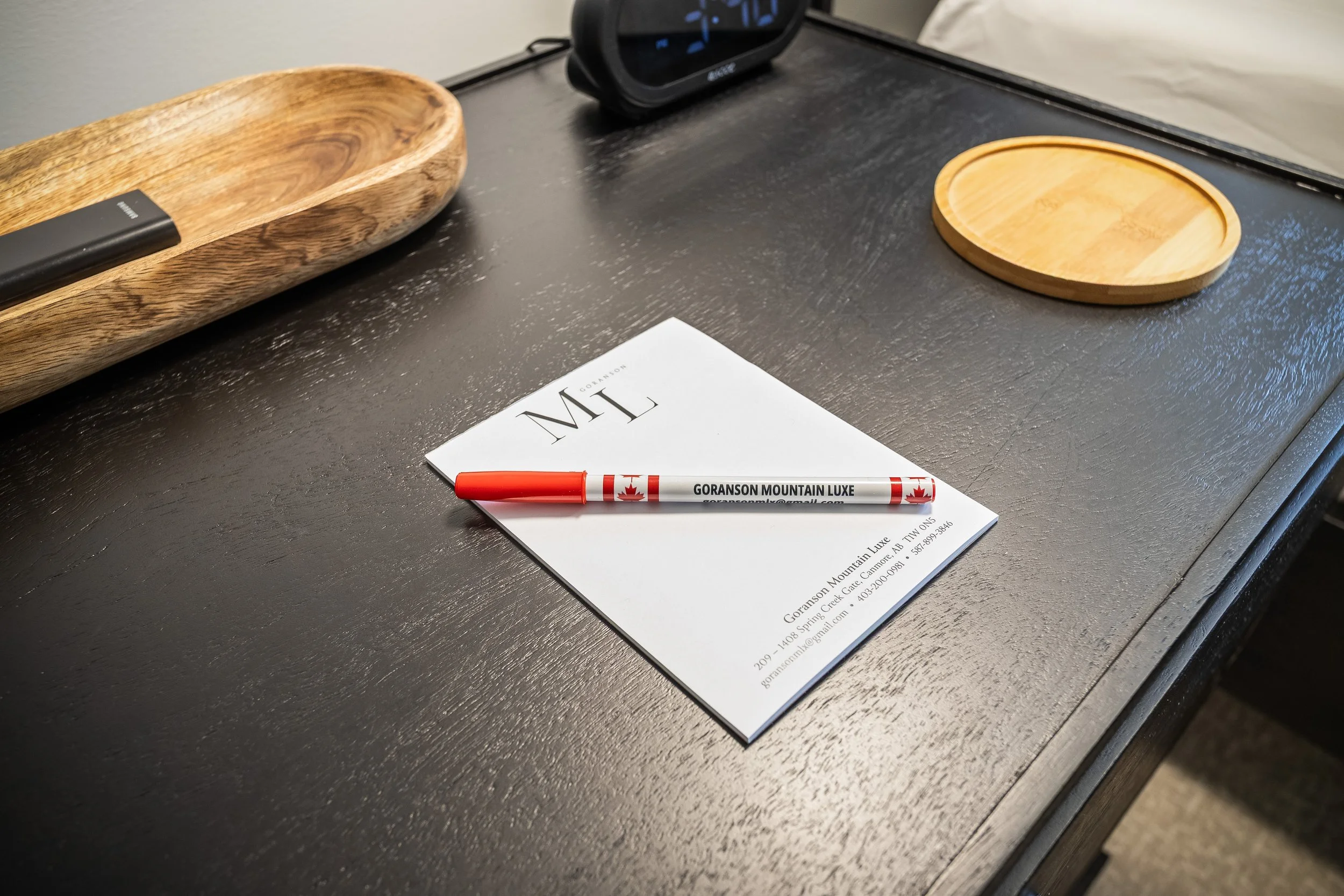 A black desk with a wooden tray, a round wooden dish, a black alarm clock, and a piece of paper with a red marker on top. The paper has text including a phone number and address for GORANSON MOUNTAIN LUXE.
