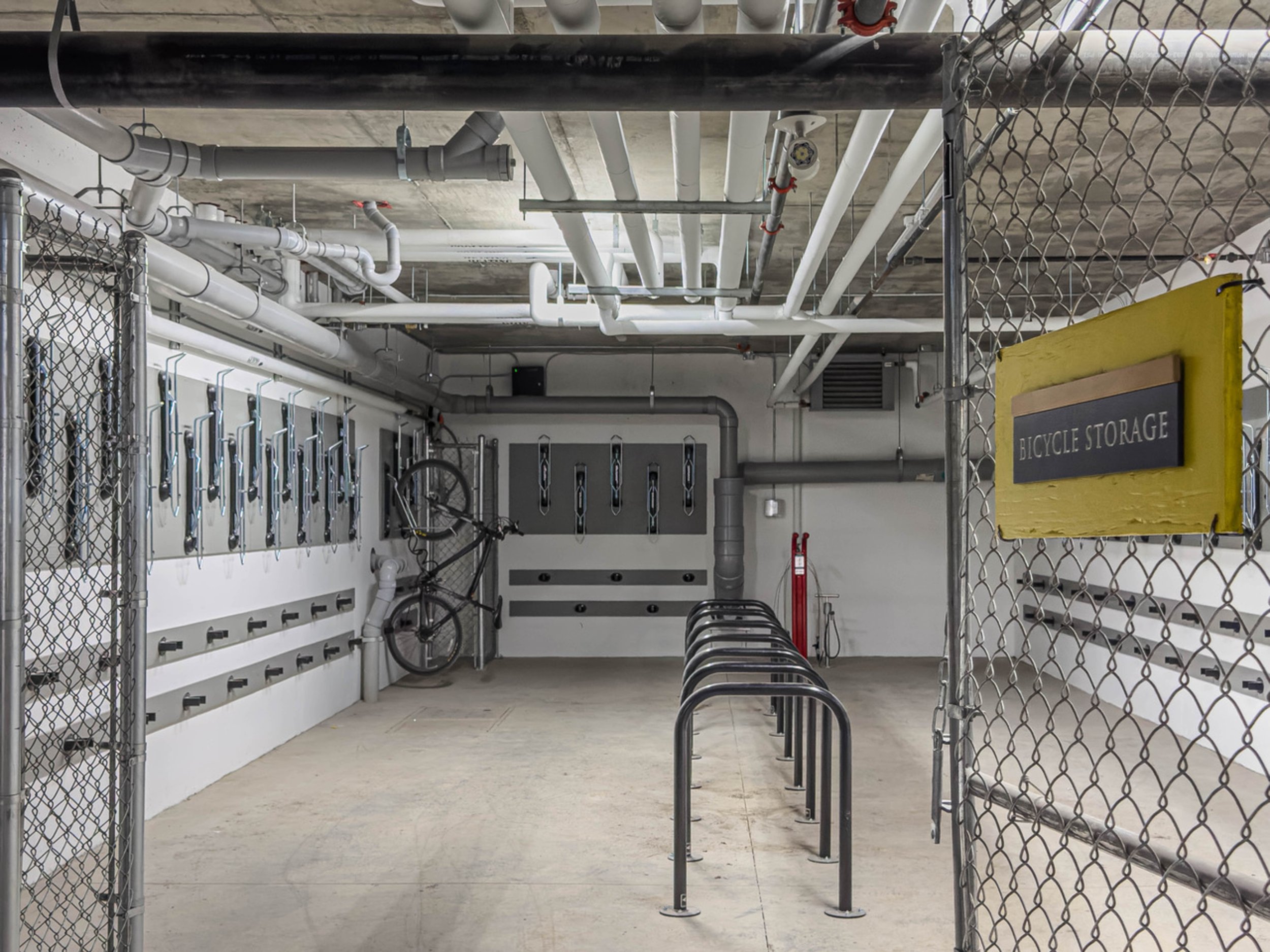 Empty bicycle storage area with a few bikes mounted on a wall, bike racks, and a yellow sign reading 'BICYCLE STORAGE' behind a chain-link fence.