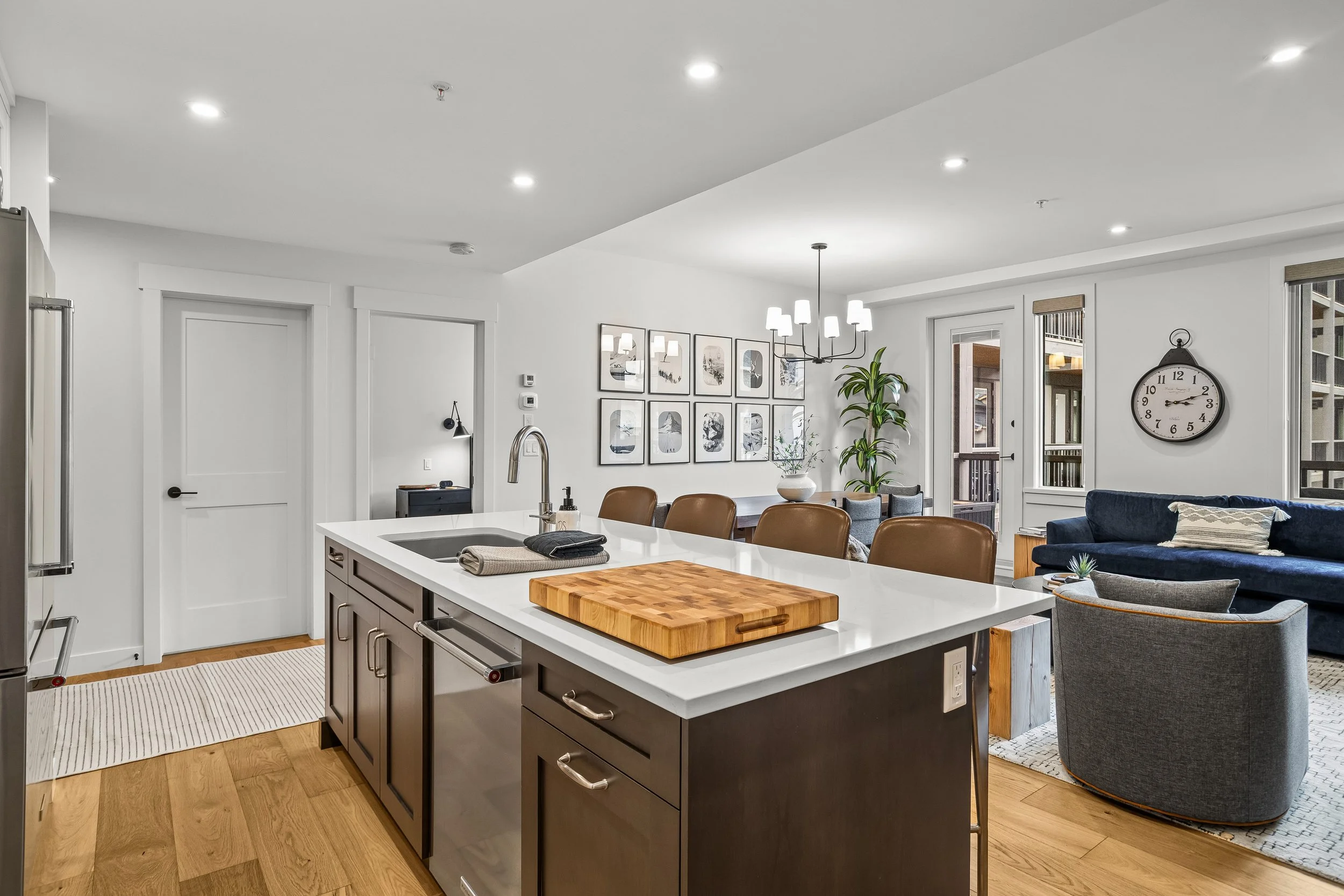 Open-concept living room and kitchen with white walls, hardwood flooring, a kitchen island with a cutting board, brown leather chairs, a dark blue sofa, a gray armchair, artwork on the wall, a large wall clock, and large windows with blinds
