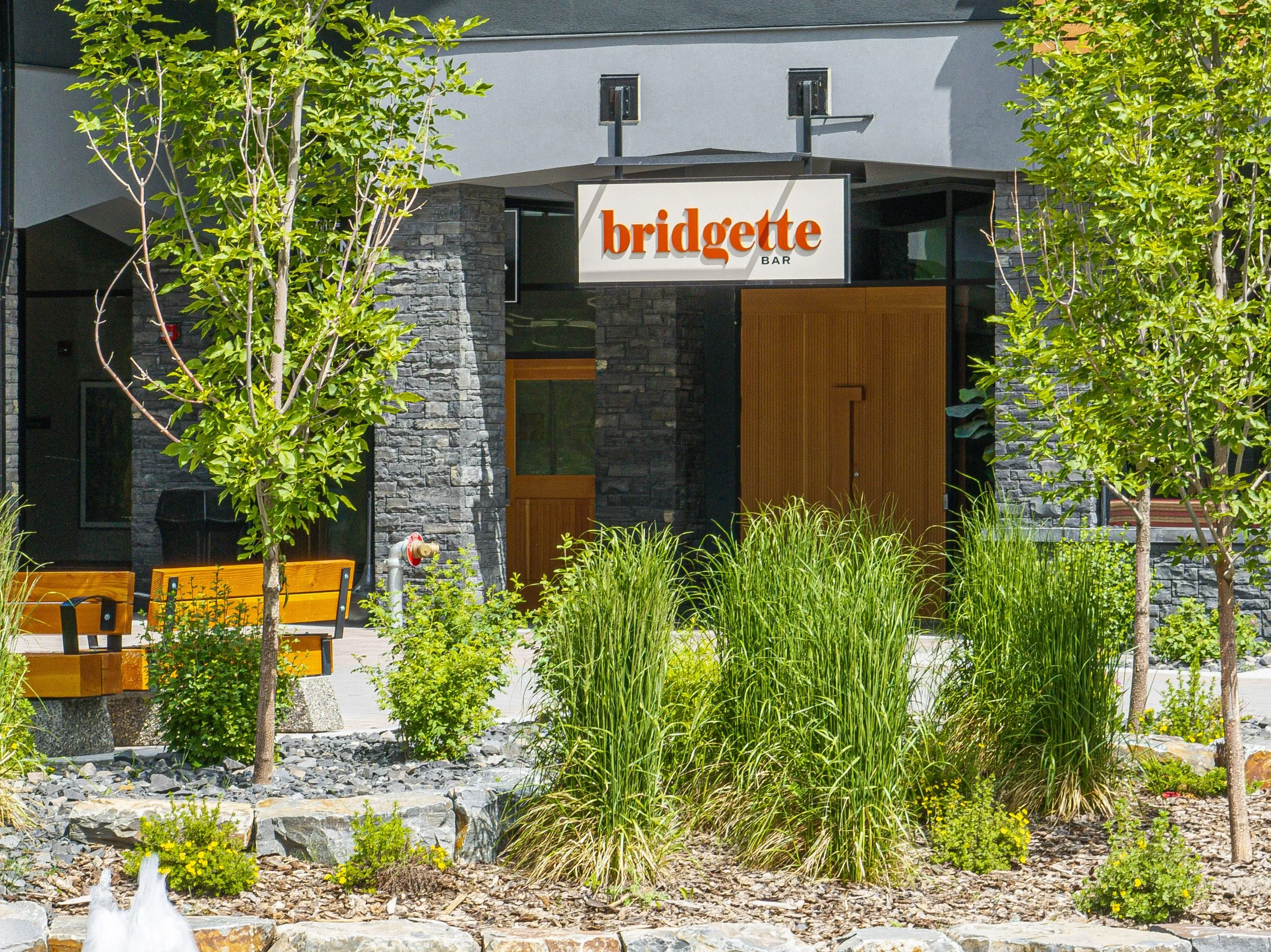 Exterior of a restaurant called Bridgette Bar with modern gray stone facade and wooden doors, surrounded by green trees, bushes, and ornamental grasses, with benches in the outdoor seating area.