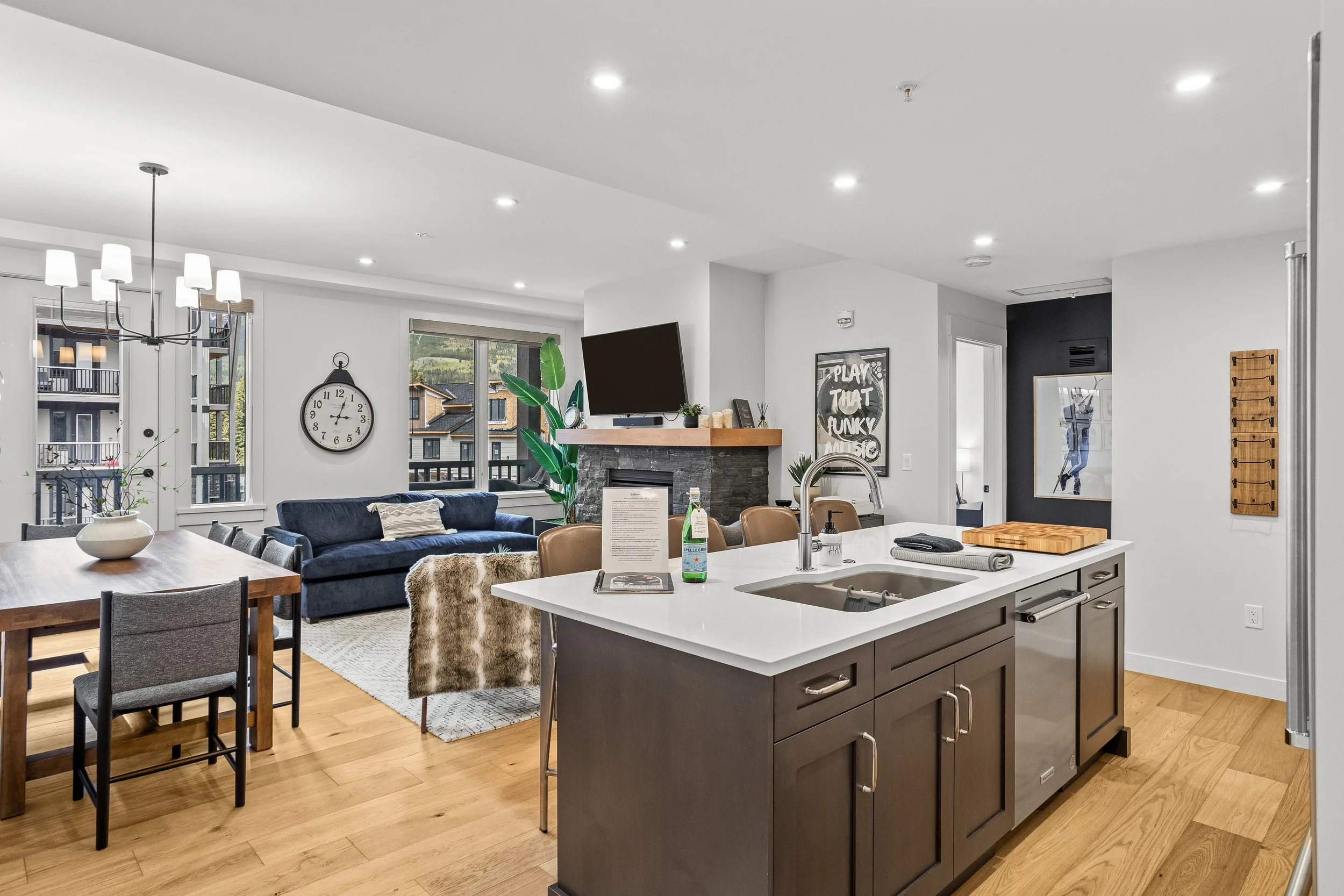 Open-concept living room and kitchen area with hardwood floors, a navy blue couch, large windows, wall clock, white walls, various decor, and a mounted TV above a fireplace.