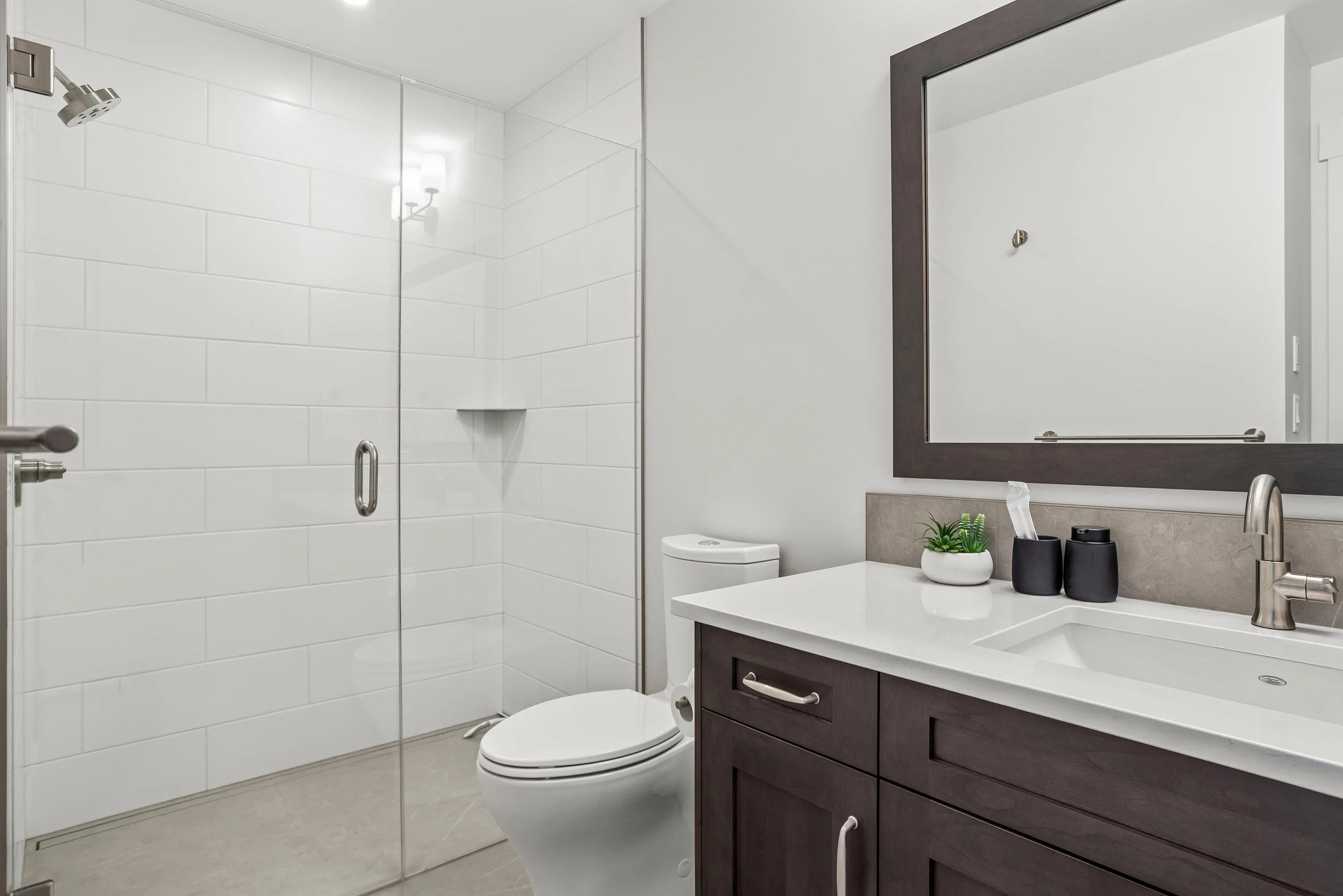 Modern bathroom with walk-in shower, white tiled walls, toilet, dark wood vanity with white countertop, large mirror, small plant, black containers, and silver fixtures.