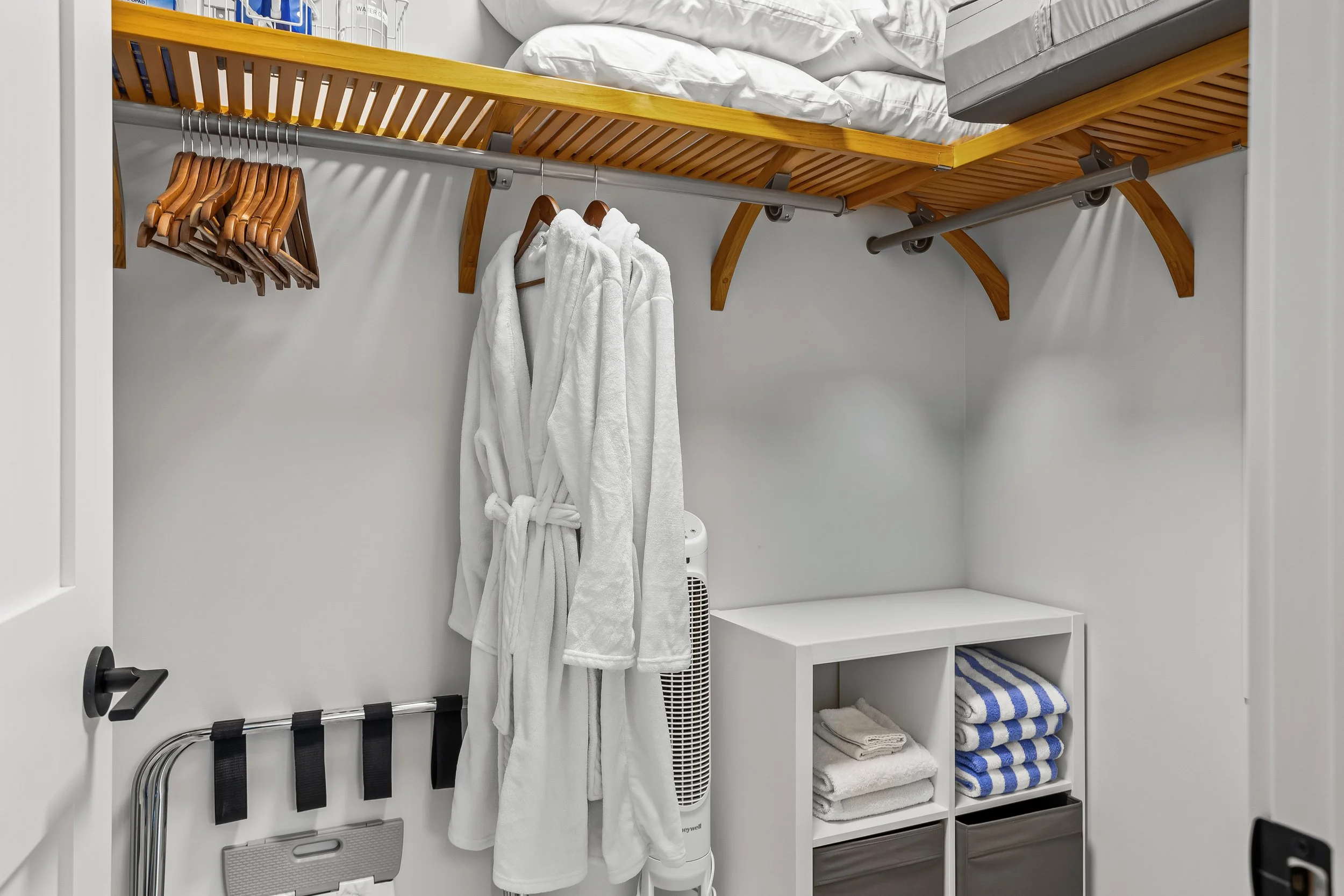 A closet with a wooden shelf and rod holding hangers, white bathrobes, a white tower fan, and a white storage cubby containing towels and bins.