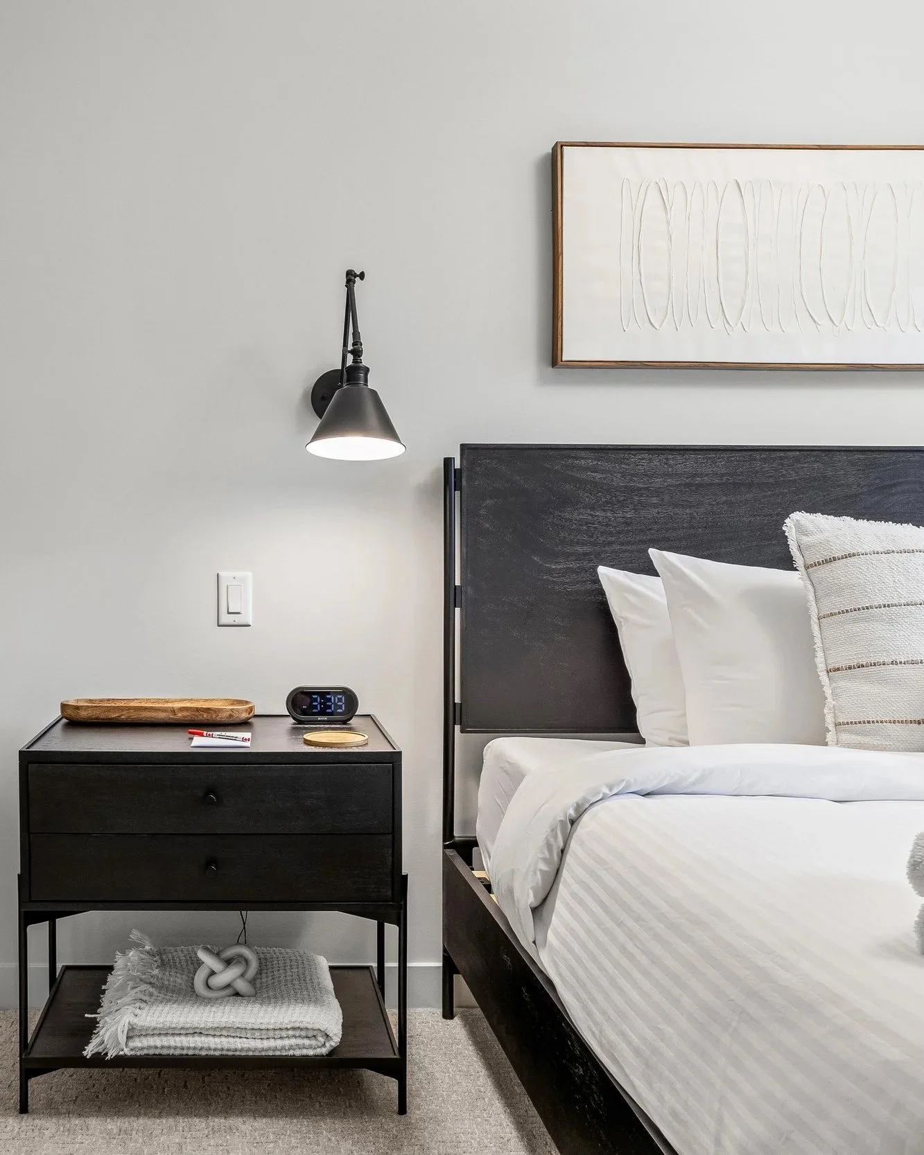 Modern bedroom with a black nightstand, wall-mounted black lamp, and a minimalist artwork above the bed. The bed has white linens and pillows. Nightstand contains a wooden tray, digital alarm clock, and a coaster, with a folded blanket and decorative knot underneath.