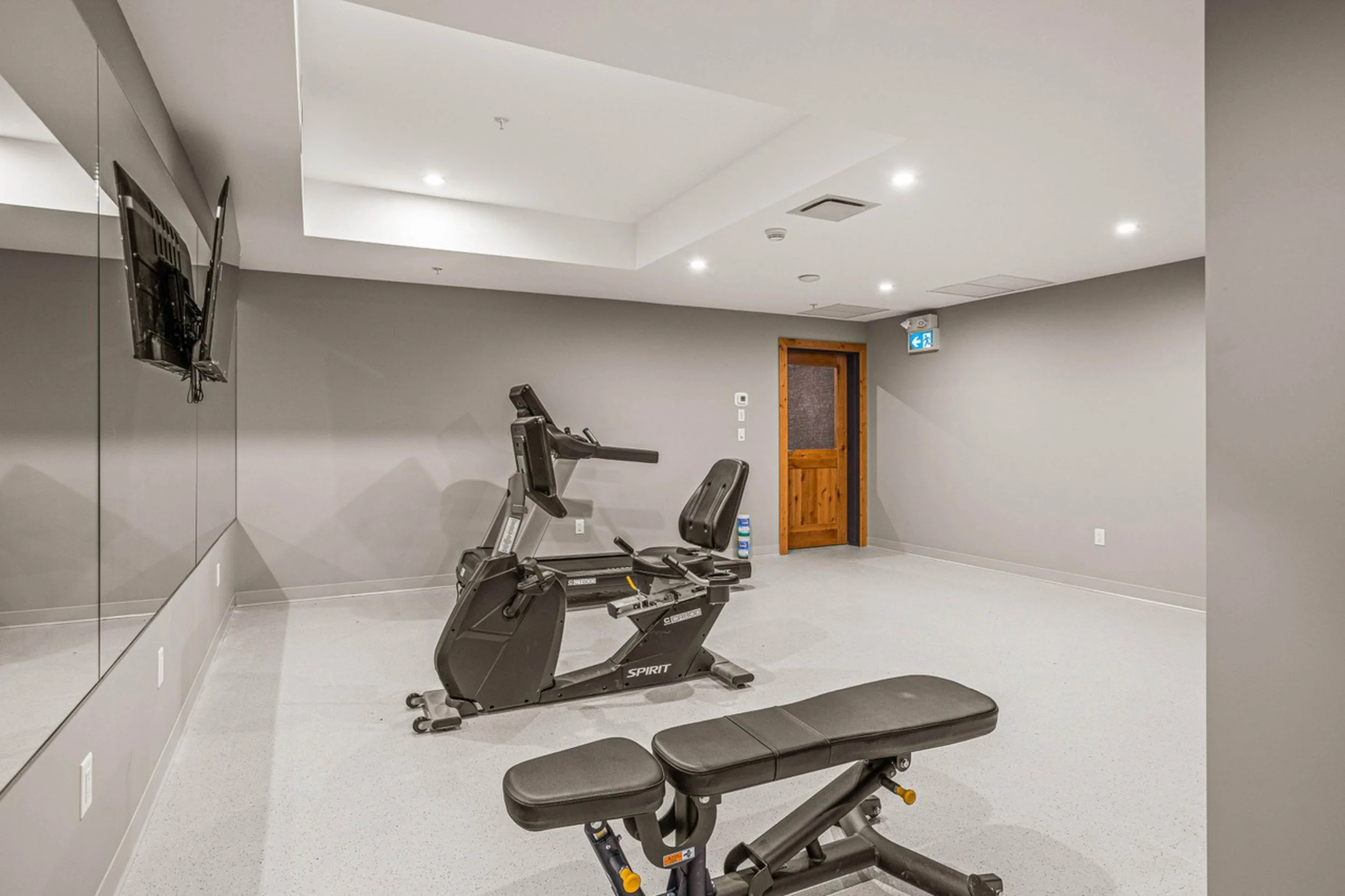 Empty indoor gym with stationary exercise bike and workout bench, gray walls, white ceiling with recessed lights, wall-mounted TV, wooden door, and an emergency exit sign.