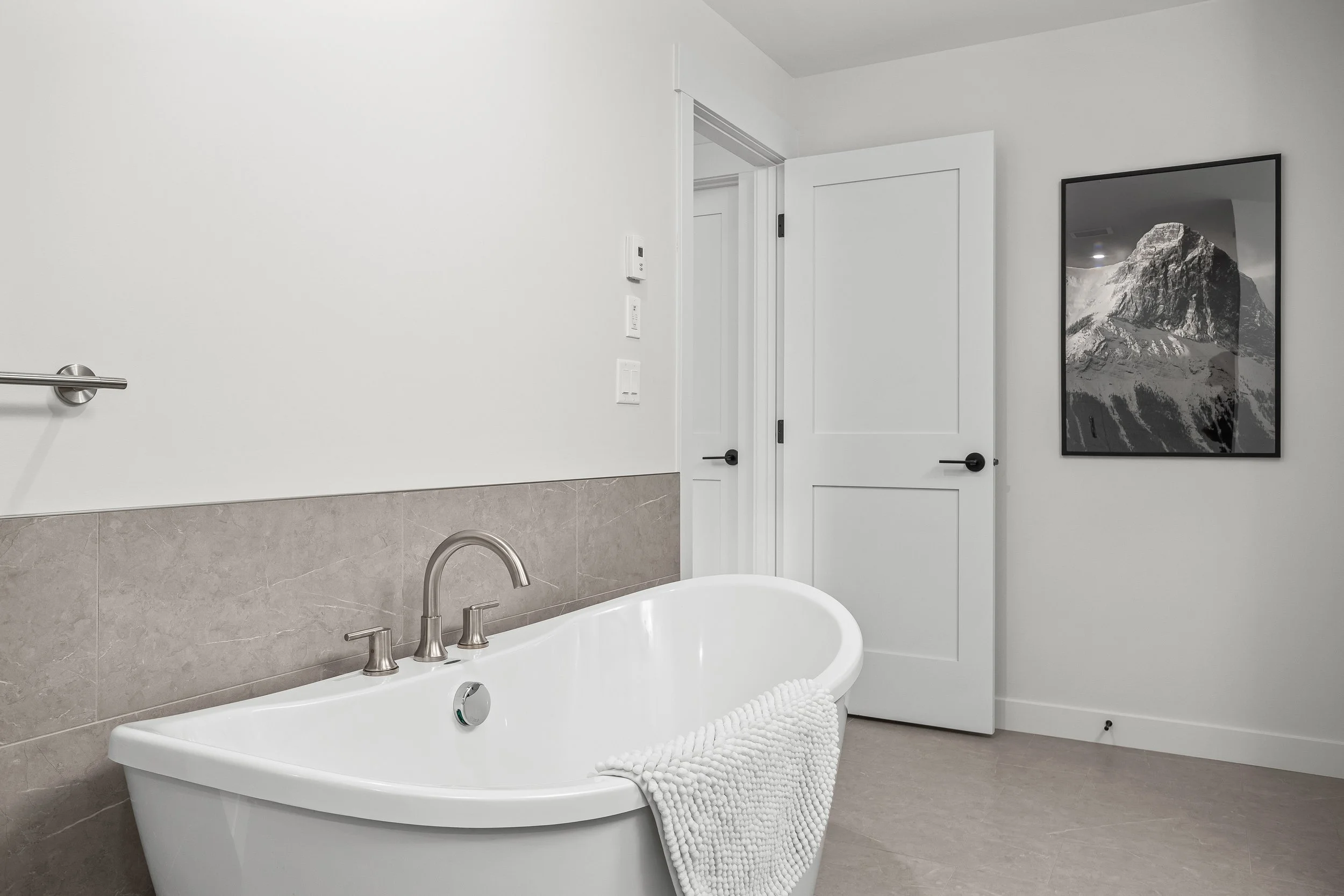 A modern bathroom with a freestanding bathtub in front of a wall with light brown tiles, a white door partially open, and a framed black-and-white mountain photograph on the wall.