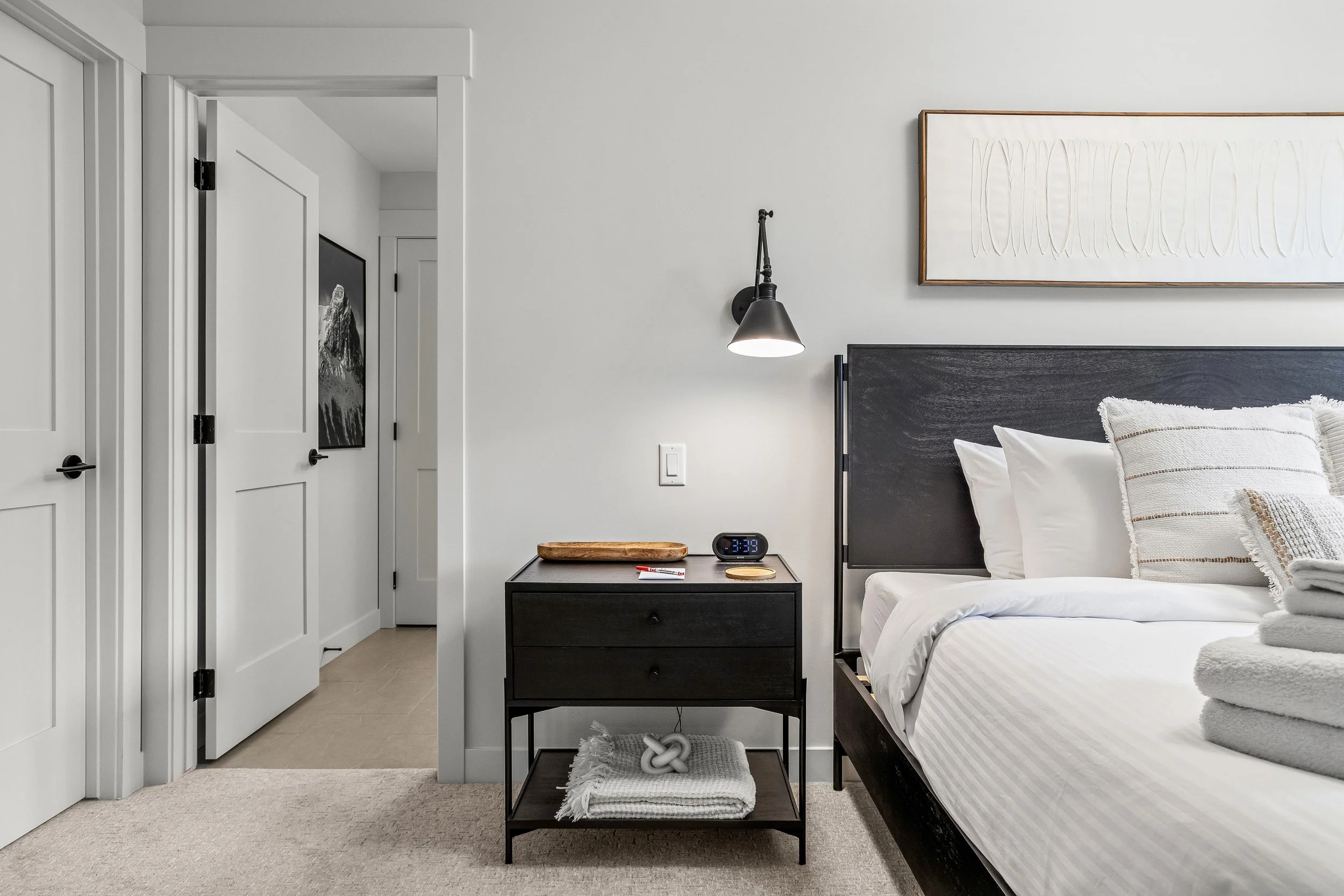 A neatly made bed with white linens and pillows in a modern bedroom. Next to the bed is a black nightstand with a digital clock, a wooden tray, and a small dish. There is a wall-mounted black lamp and abstract artwork above the bed. The room has whit