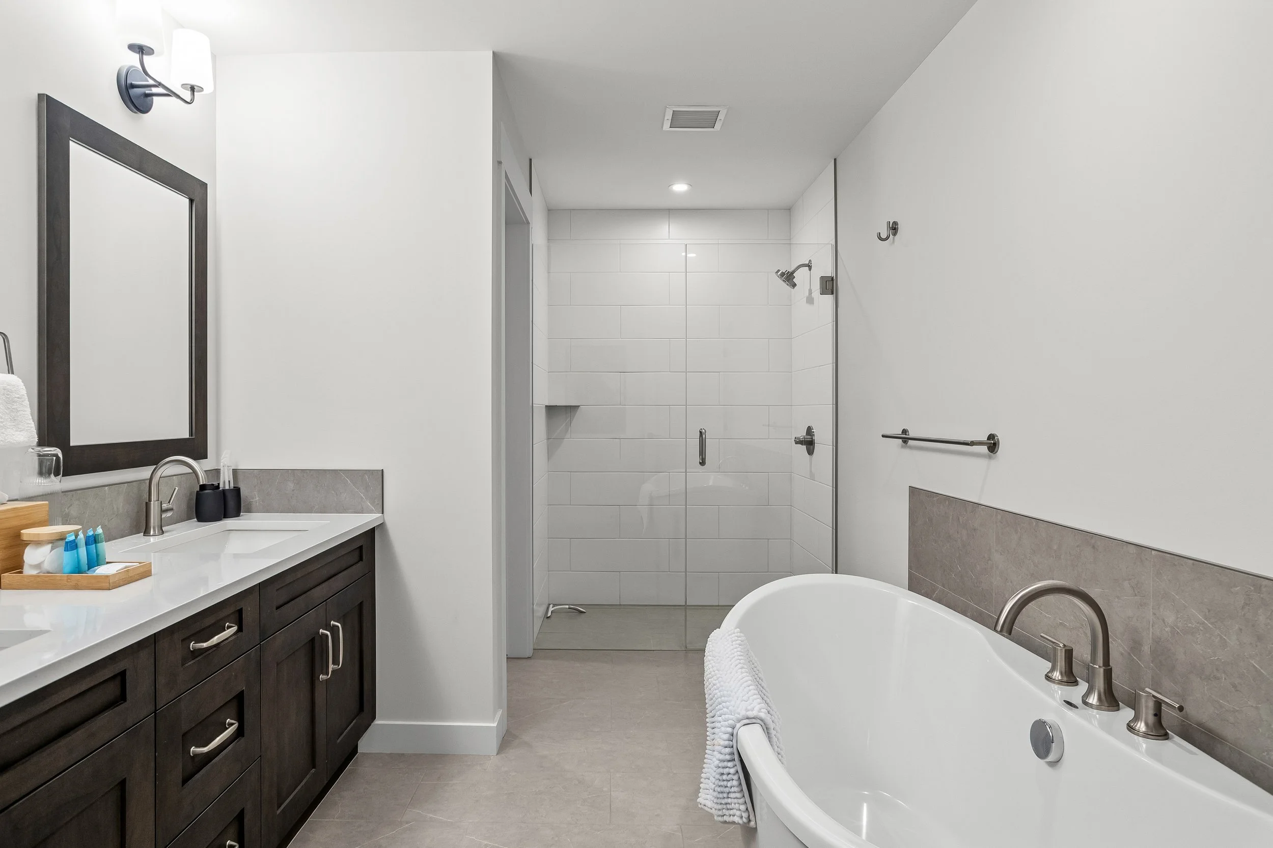 Modern bathroom with dark wood vanity, white countertop, mirror, towel rack, bathtub, glass shower enclosure, wall hooks, and neutral-colored floor and walls.