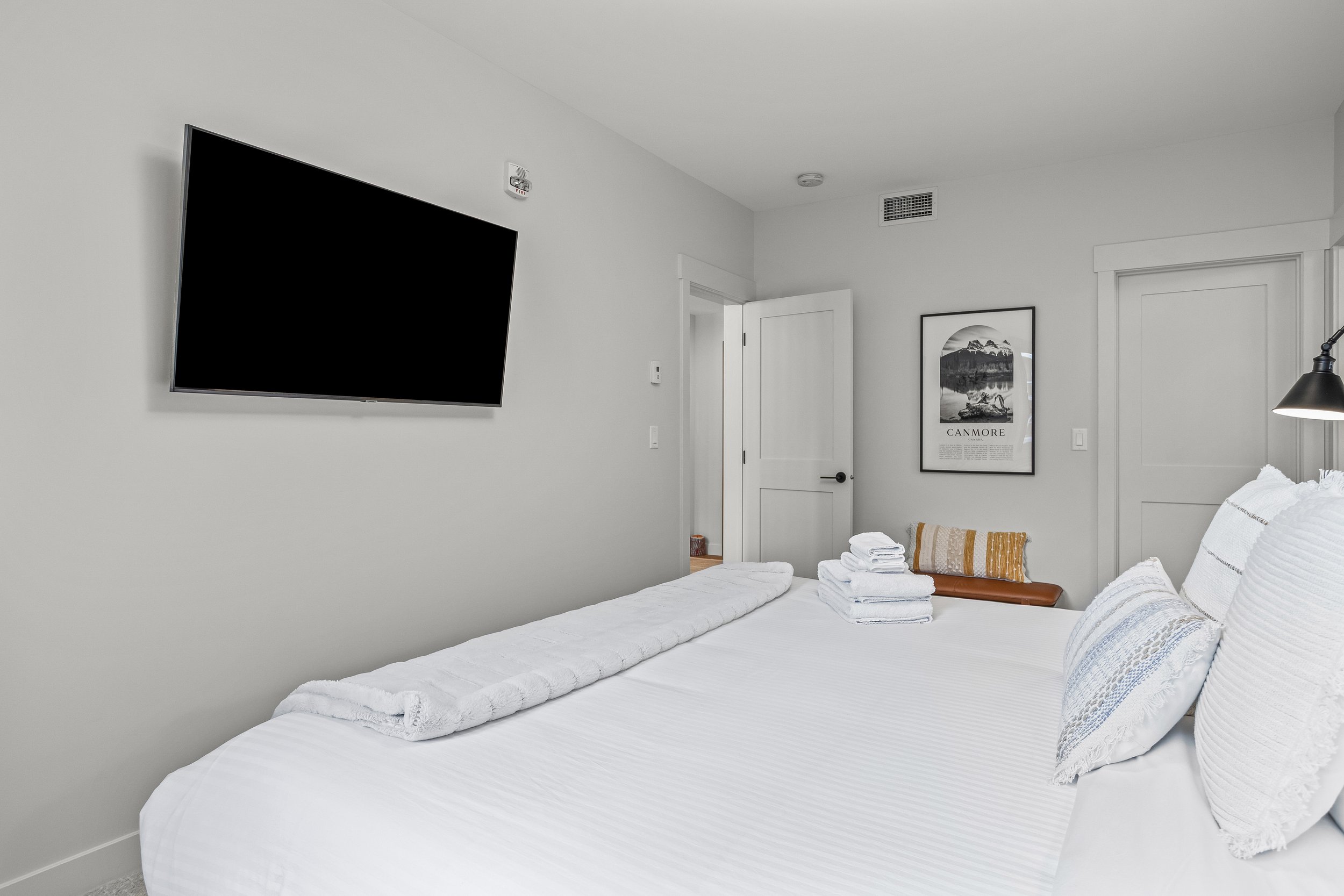 A neatly made bed with white linens and pillows in a modern minimalistic bedroom. Towels are folded on the bed, and a framed art print titled 'Canmore' hangs on the wall. There is a mounted flat-screen TV on the wall opposite the bed, and a door lead