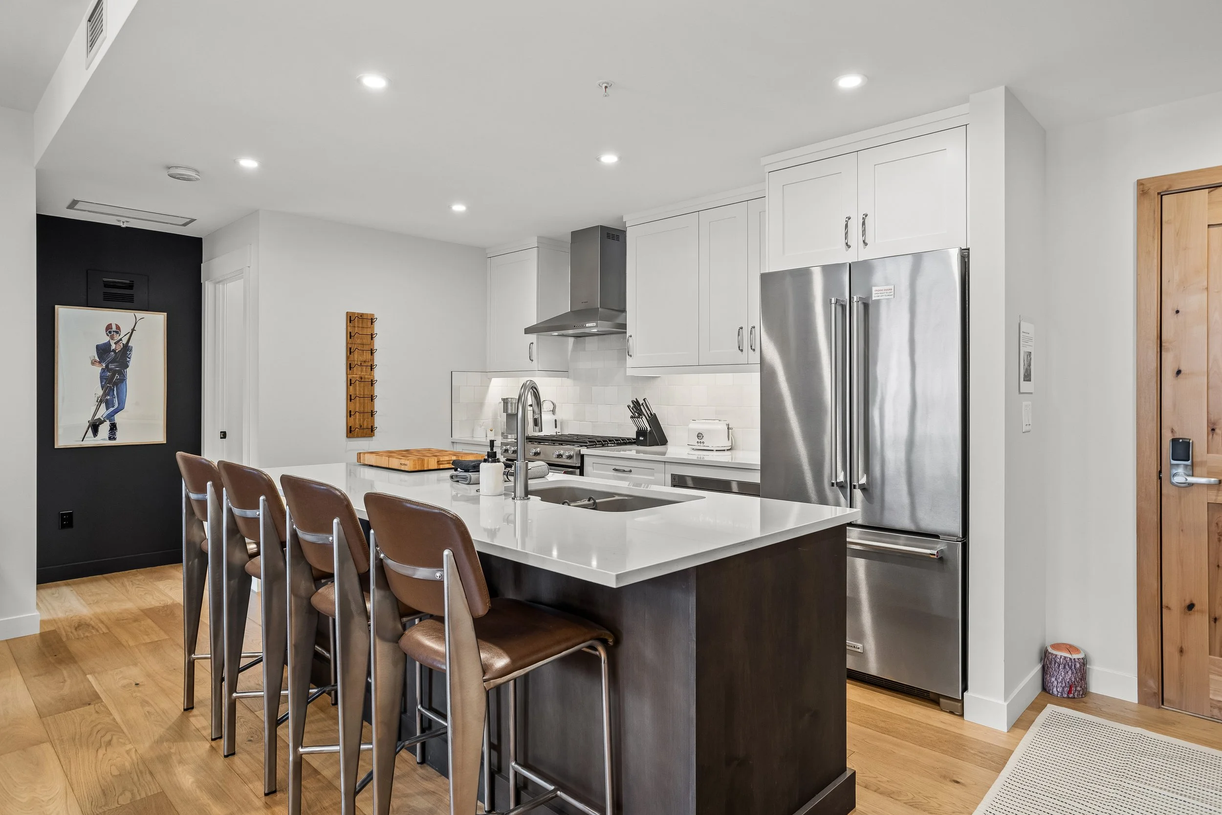 Modern kitchen with white cabinets, stainless steel refrigerator, kitchen island with bar stools, black accent wall, wooden floor, and minimal decor.