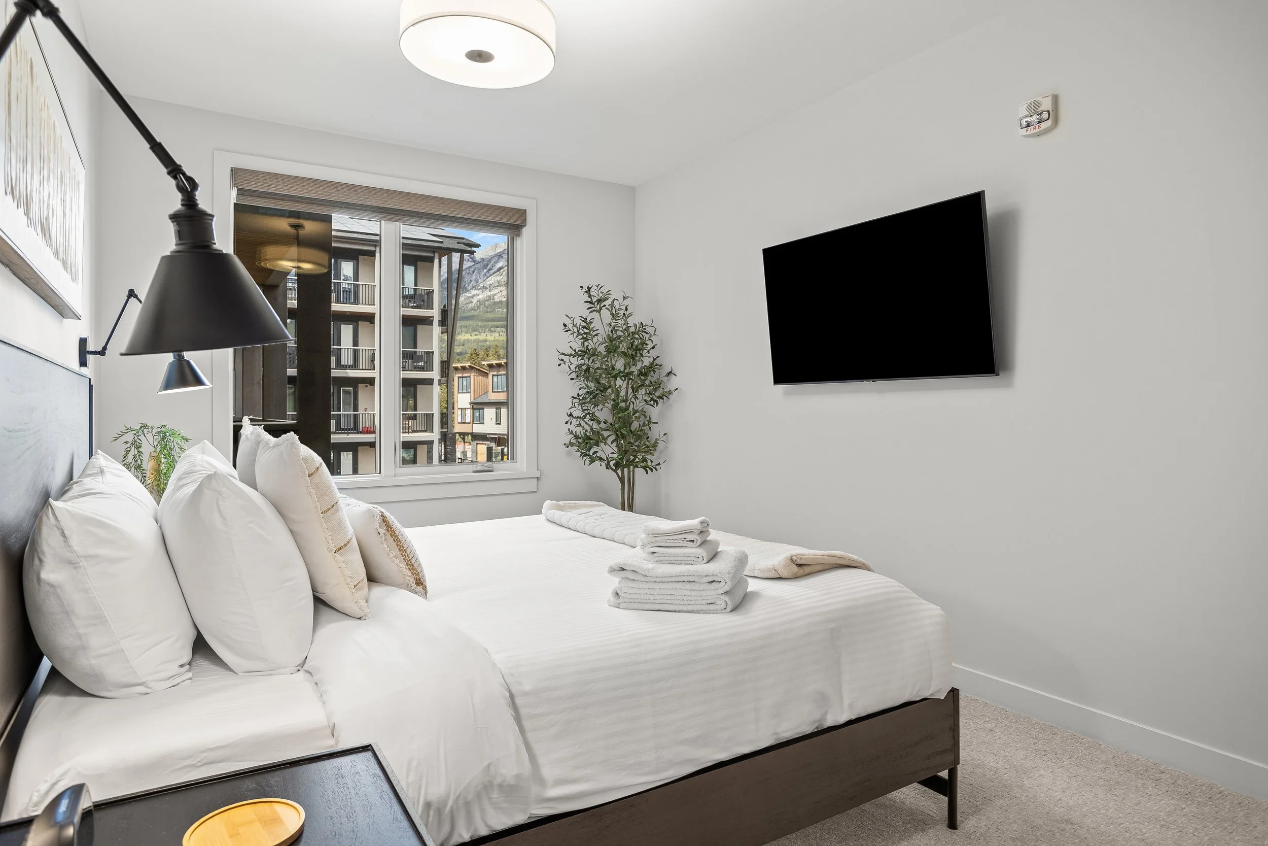 A bedroom with a bed, stacked towels, window overlooking a building and mountains, wall-mounted TV, plant, and modern decor.