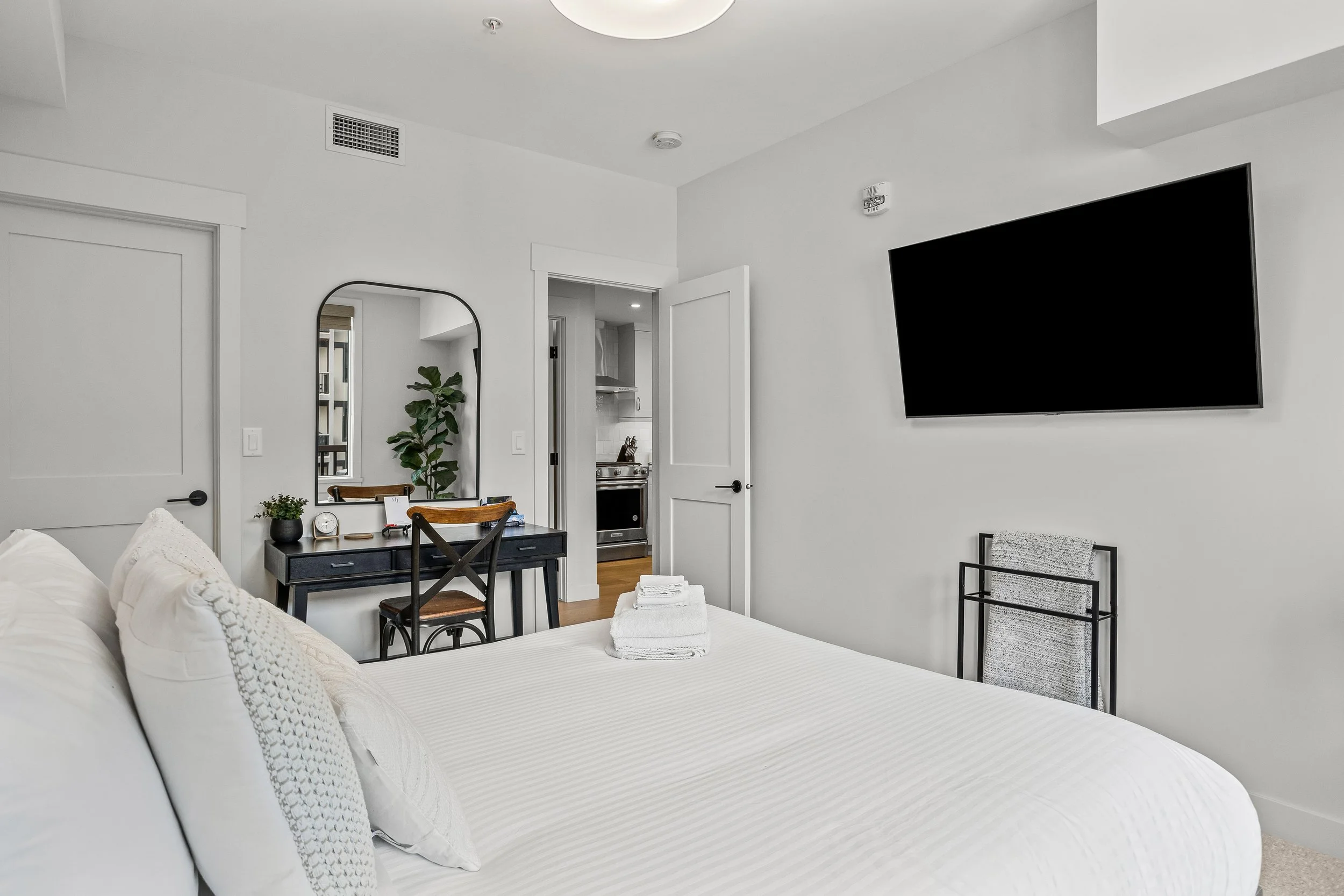 A bedroom with white walls and bedding, a wall-mounted TV, a mirror, a desk with a chair, and an open door leading to a kitchen area.