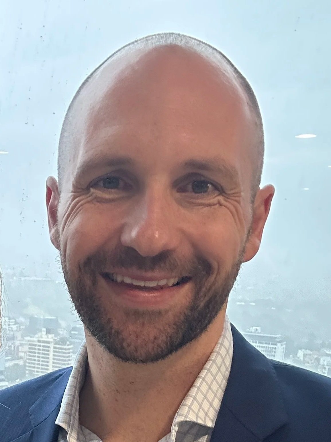 A smiling man with a beard and a shaved head, wearing a checkered shirt and a blazer, standing in front of a window overlooking a cityscape.