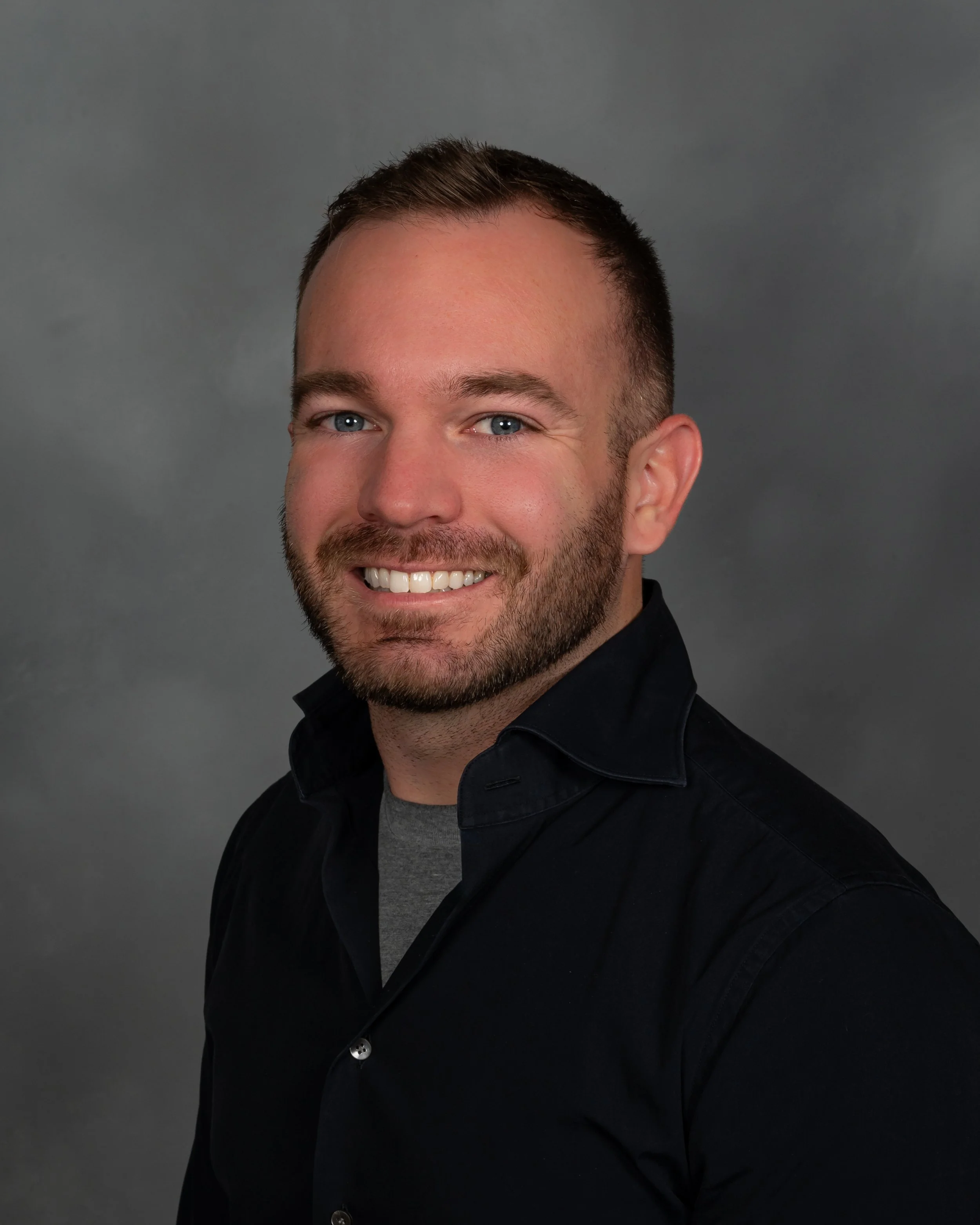 Headshot of a man with short brown hair, a beard, blue eyes, wearing a black button-up shirt and a gray T-shirt in front of a gray background. The man is Derek Herman, a communications executive, specializing in internal and external communications.